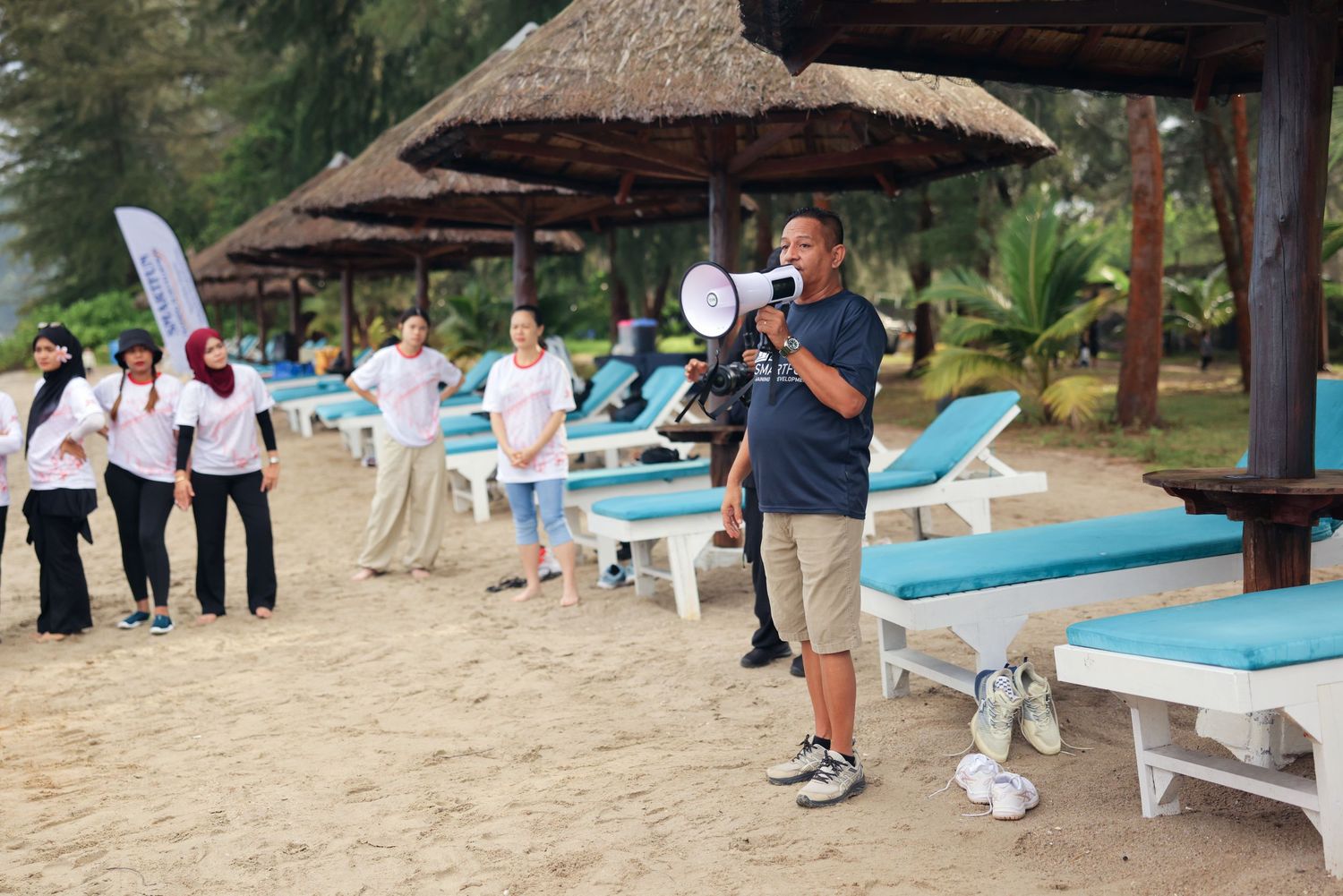 Man with megaphone addresses group on sandy beach resort with thatched umbrellas and turquoise lounge chairs.