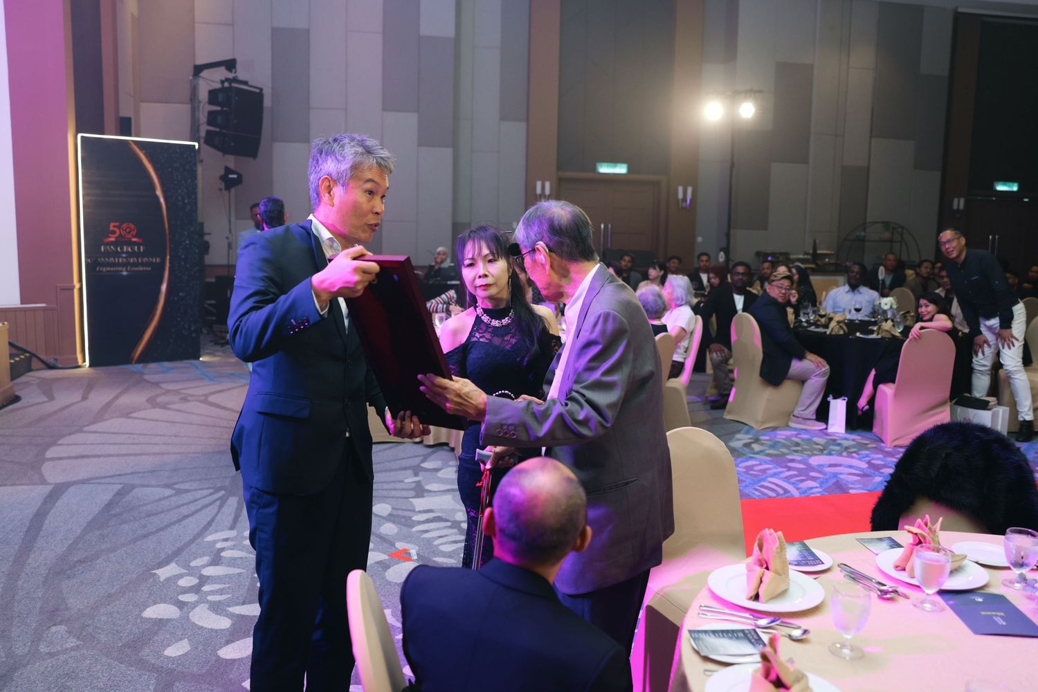 Two men exchanging an award plaque at a formal corporate gala dinner ceremony in ballroom.