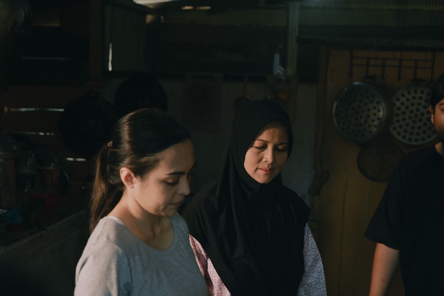 Two people stand in a dimly lit kitchen with hanging pots visible in the background.