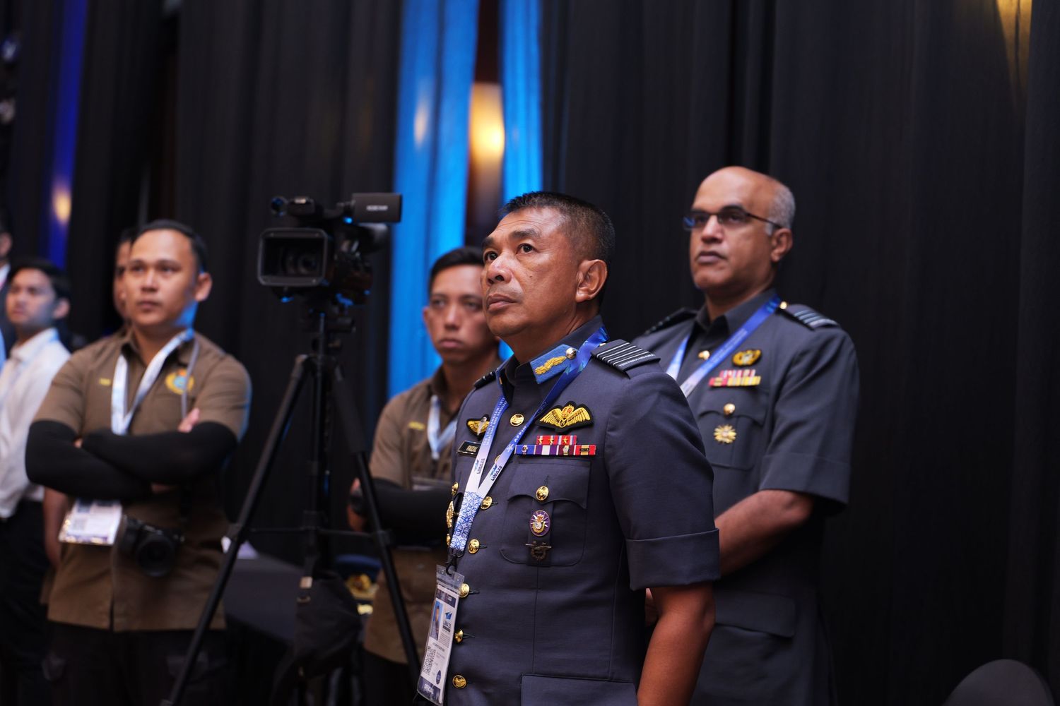Officers in military dress uniforms attend a professional conference in a dimly lit venue.