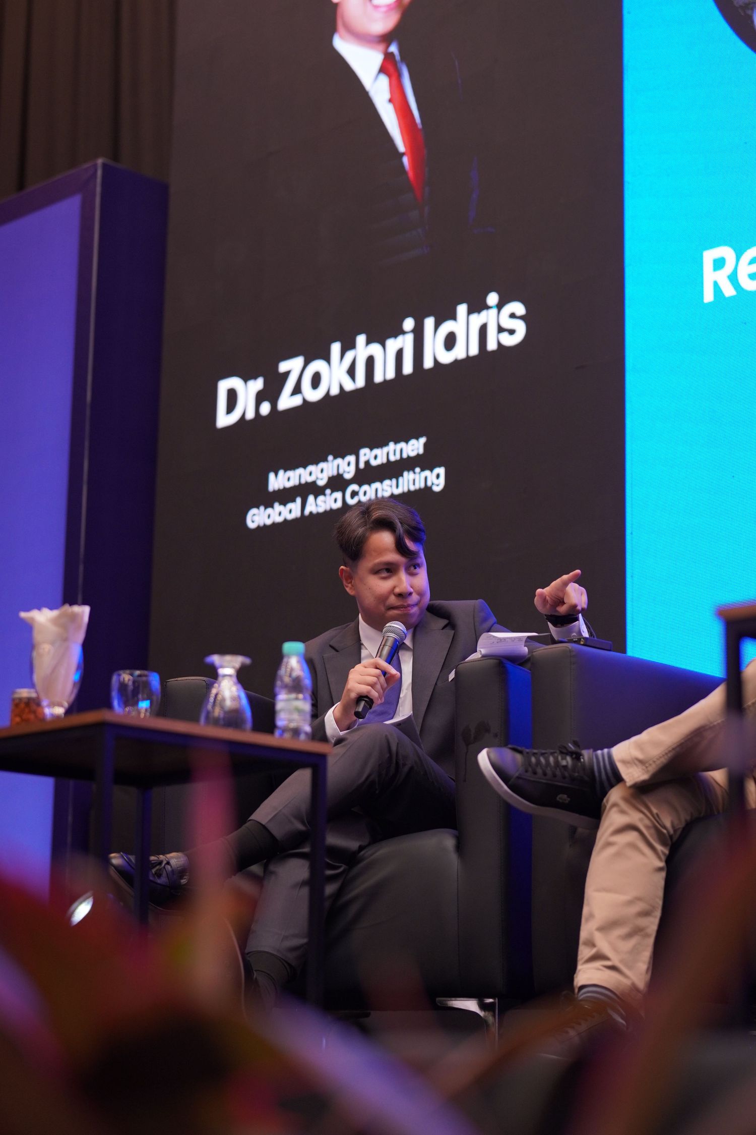 Speaker addresses audience at business conference with Dr. Zokhri Idris credentials displayed on screen behind.