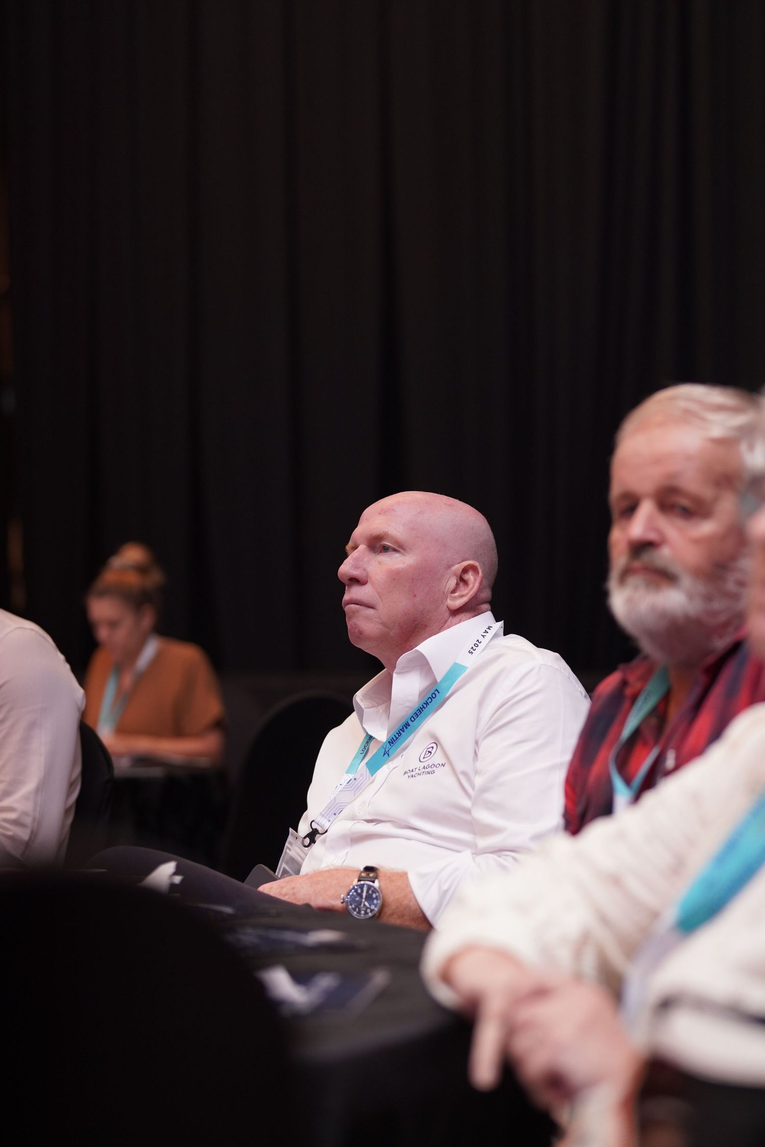 Conference attendees in white and red attire seated in dark auditorium during presentation.