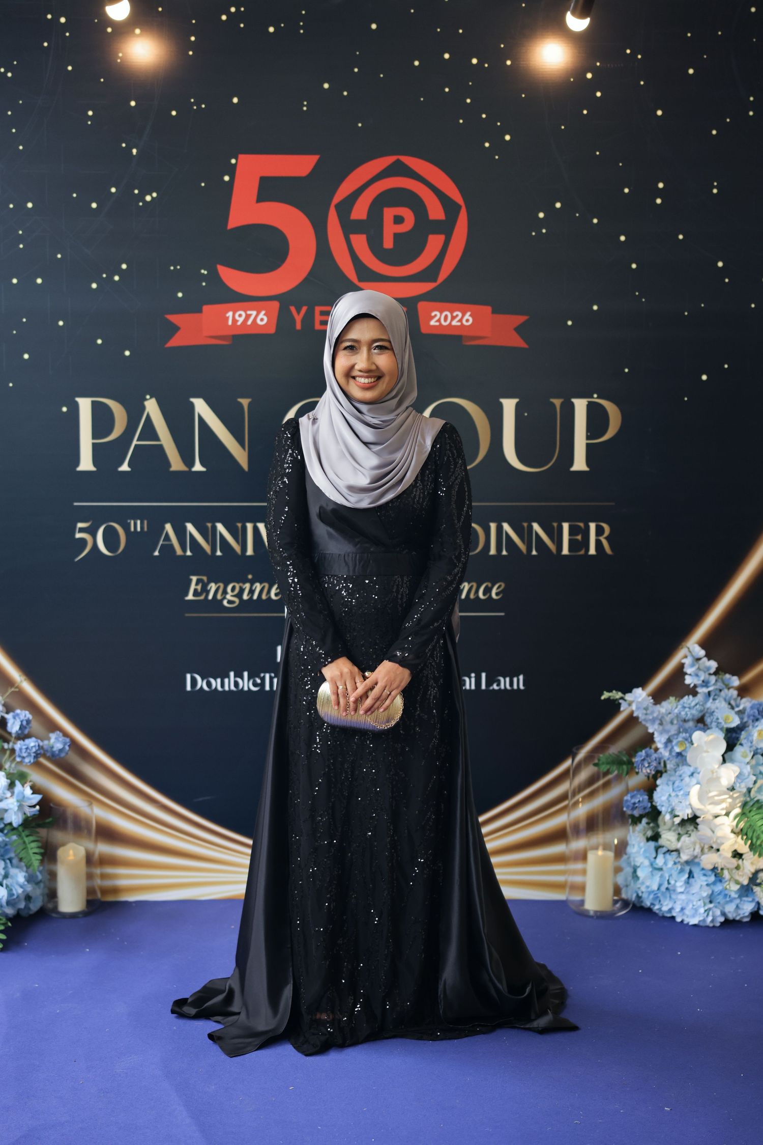 Woman in elegant black hijab dress posing at Pan Group 50th Anniversary Dinner gala event backdrop.