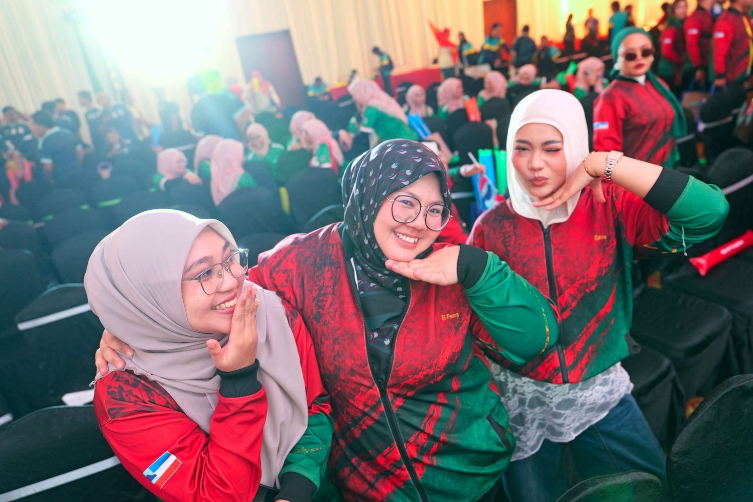 Three women in hijabs and red-green outfits pose playfully at a lively indoor event.