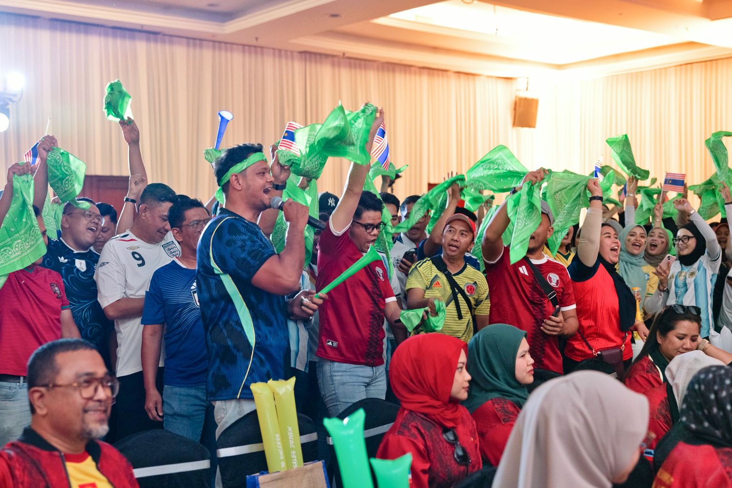 Excited crowd waving green flags and accessories at a lively indoor corporate event in Malaysia.