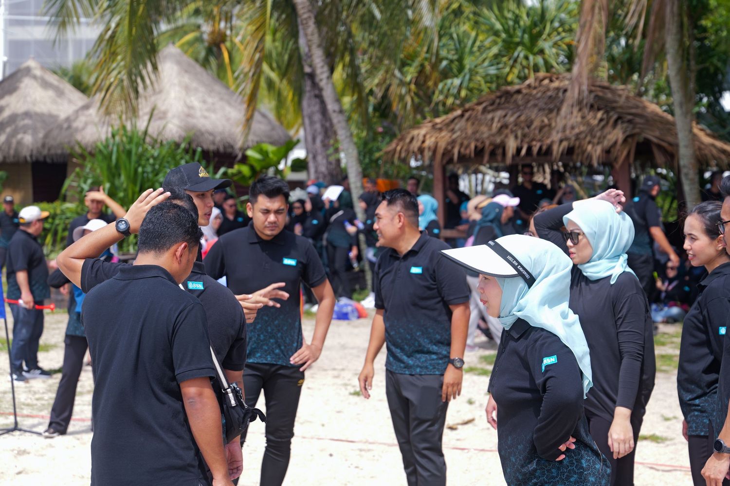 Group of people in matching black uniforms gathered on a tropical beach with palm trees and thatched huts.