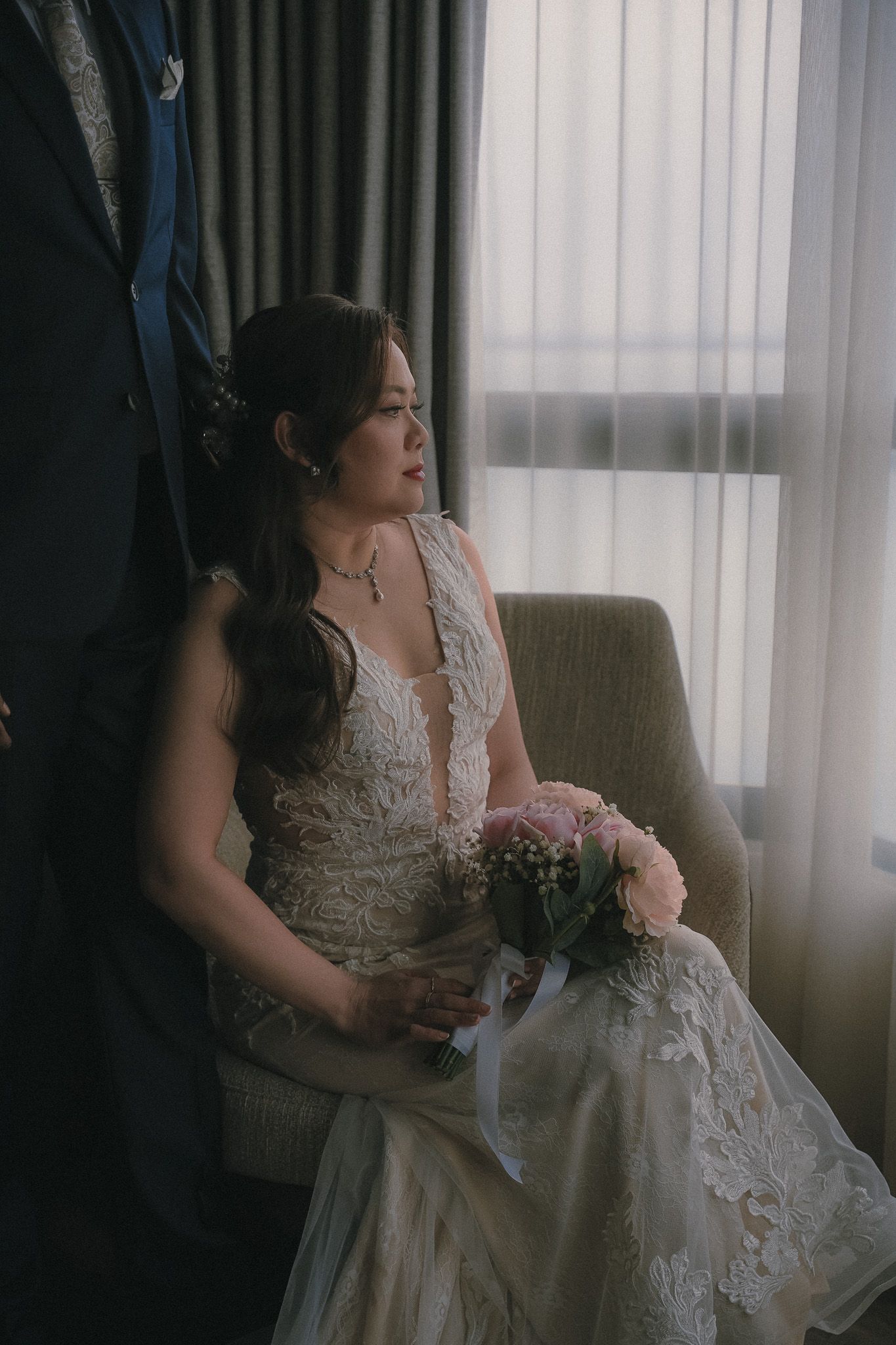 Bride in delicate lace wedding dress sits pensively by window holding pink floral bouquet in moody lighting.