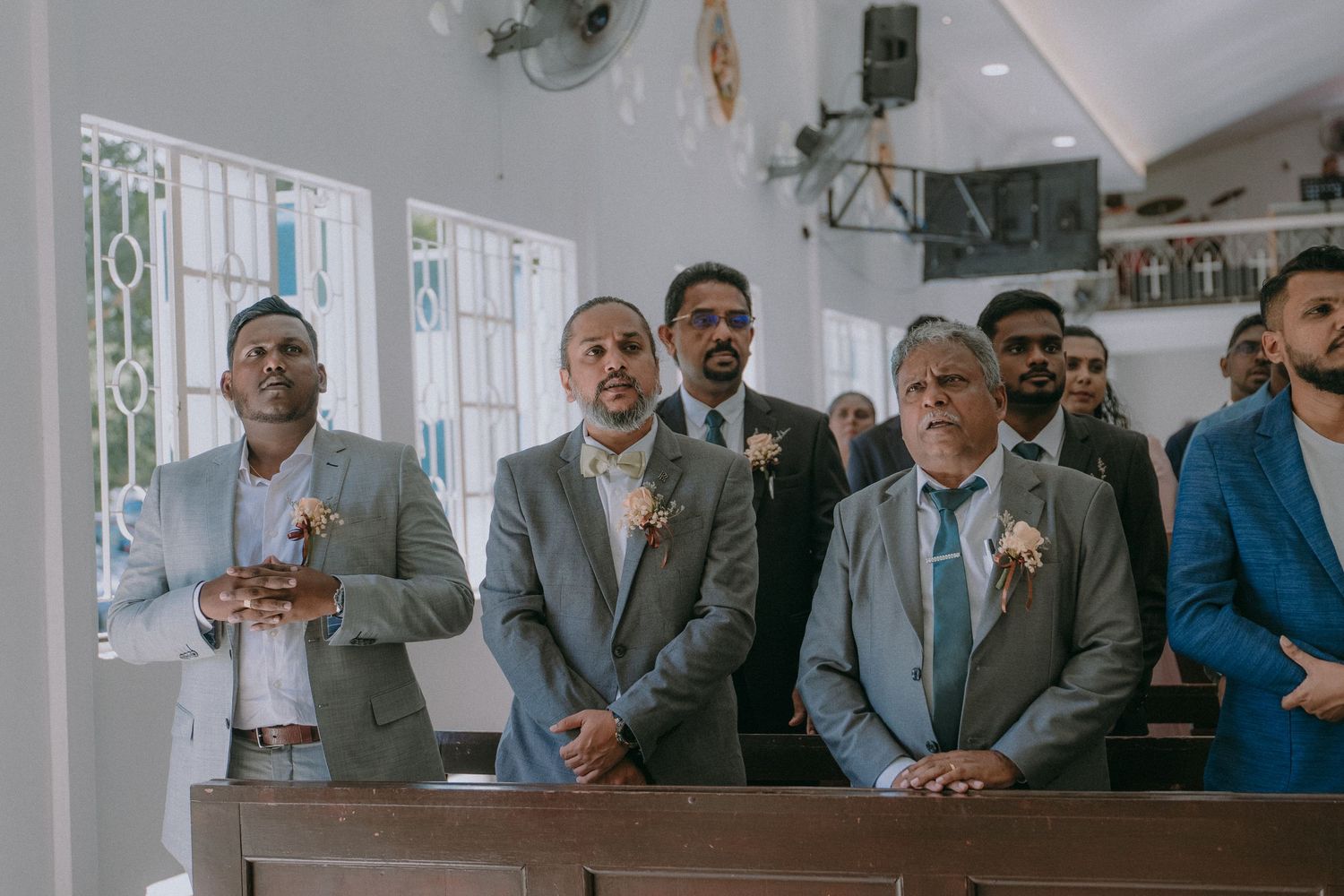 Wedding guests watch intently during a ceremony in a church with white walls and decorative windows.