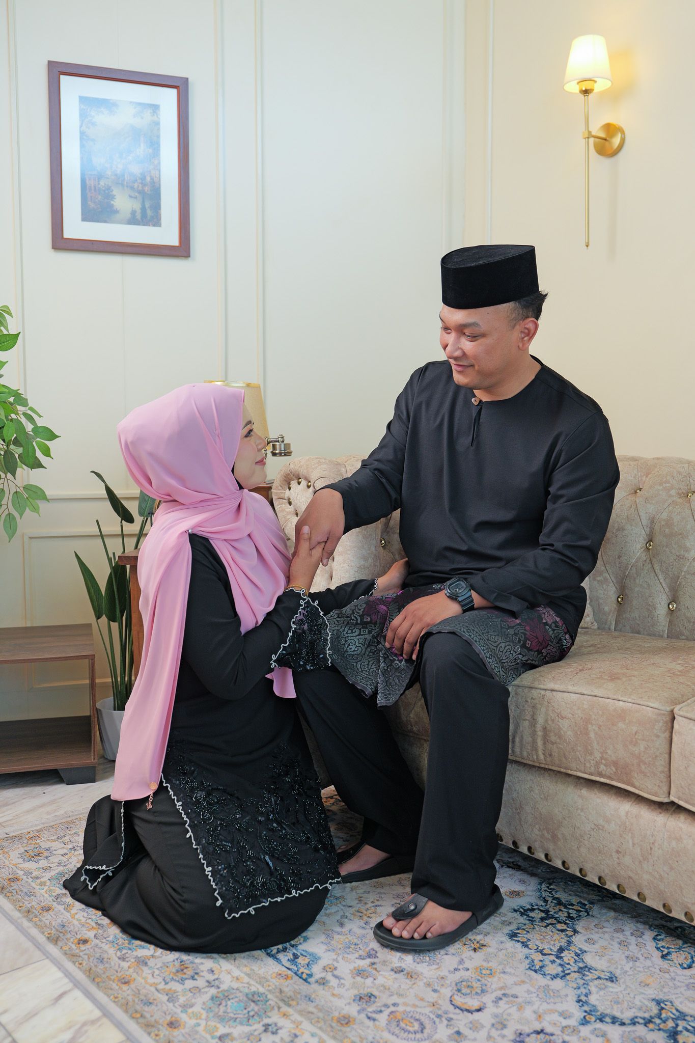 Two people in traditional black and pink attire sit together on a beige couch in an elegant room with wall sconces.