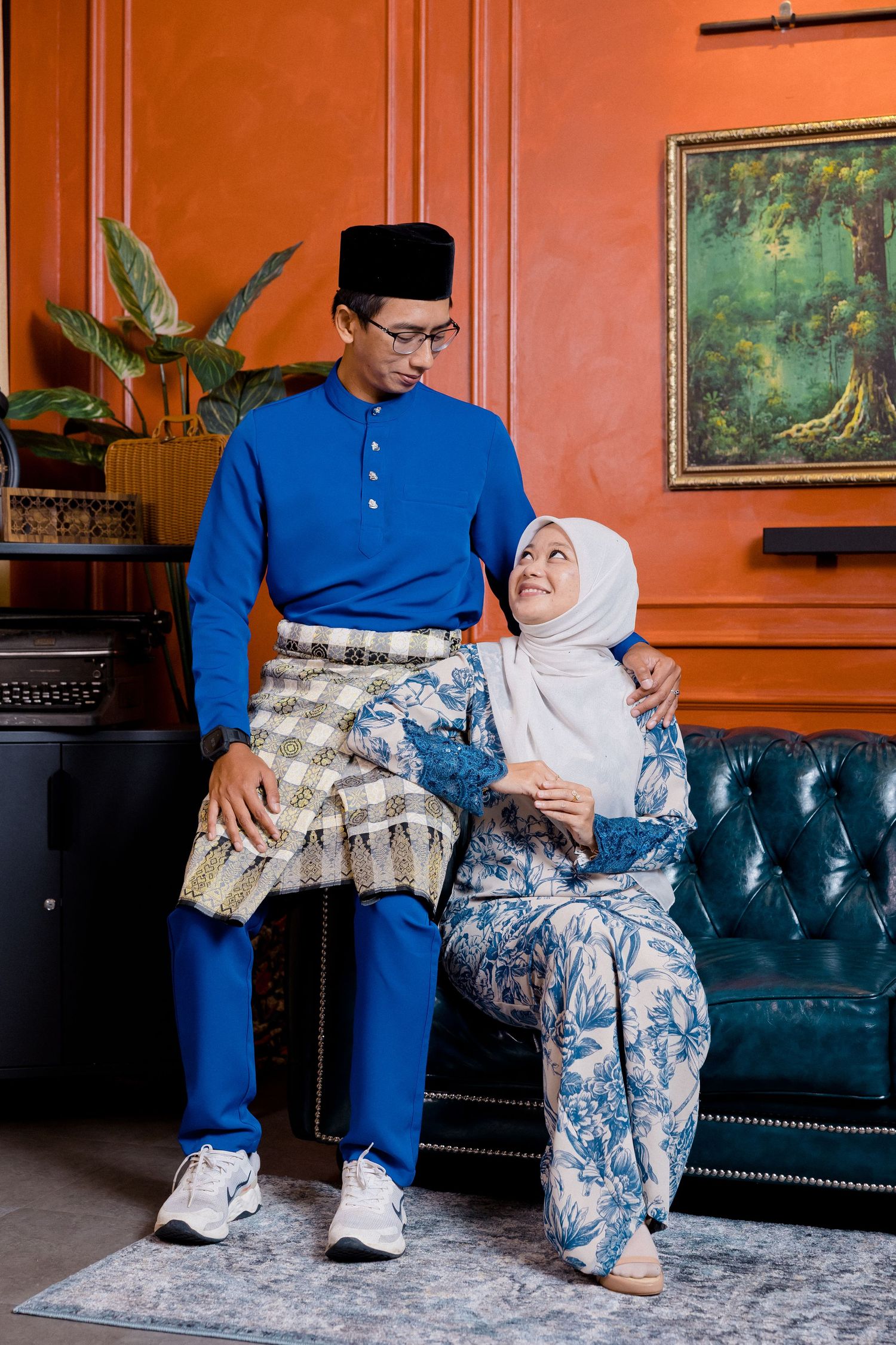 Couple in matching blue outfits and traditional attire sit together on a teal velvet couch against coral-colored walls.