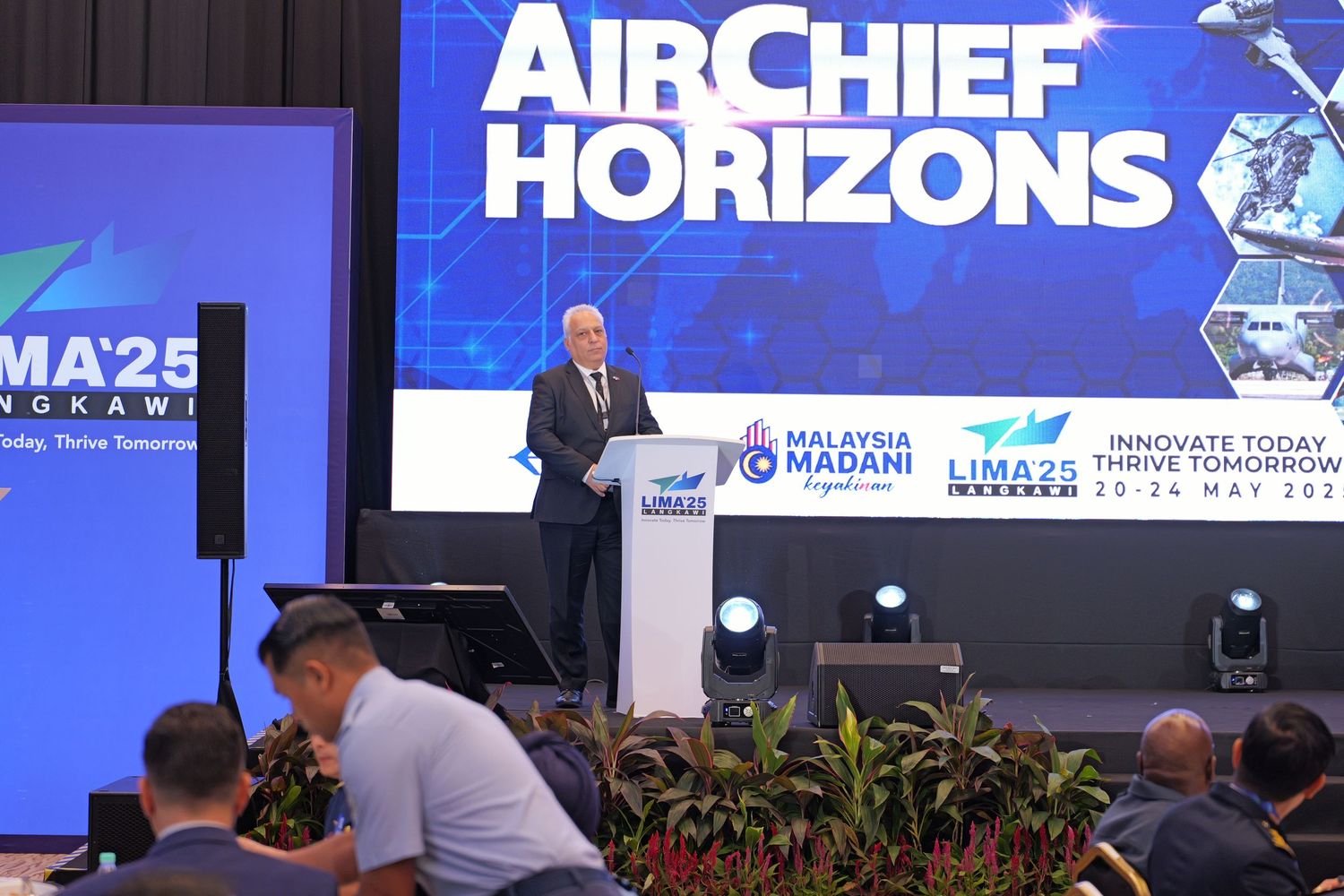Speaker presenting at AirChief Horizons conference with digital backdrop and stage lighting.