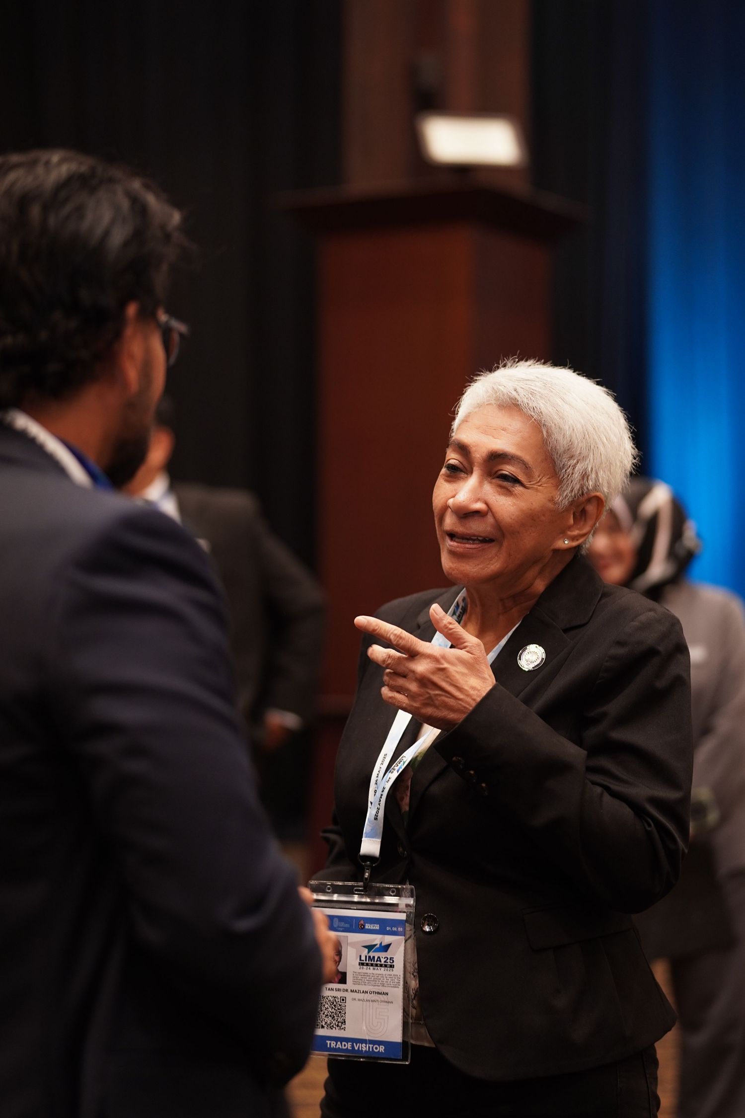 Senior attendee in black suit smiling and gesturing during a professional conference event.
