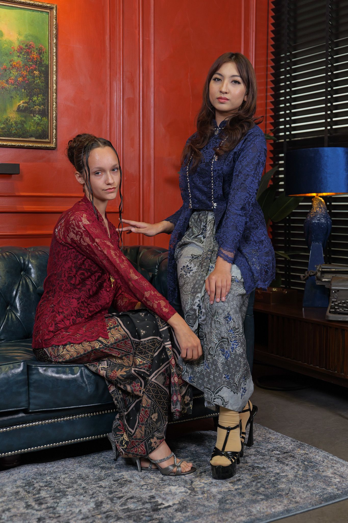 Two people pose in elegant vintage-inspired outfits with burgundy and navy lace dresses on a leather couch against red walls.