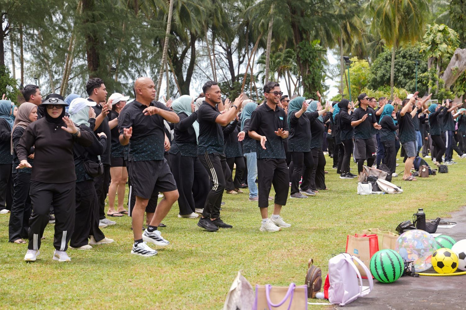 Large group of people in black uniforms performing outdoor exercise activities on a tropical beachside lawn.