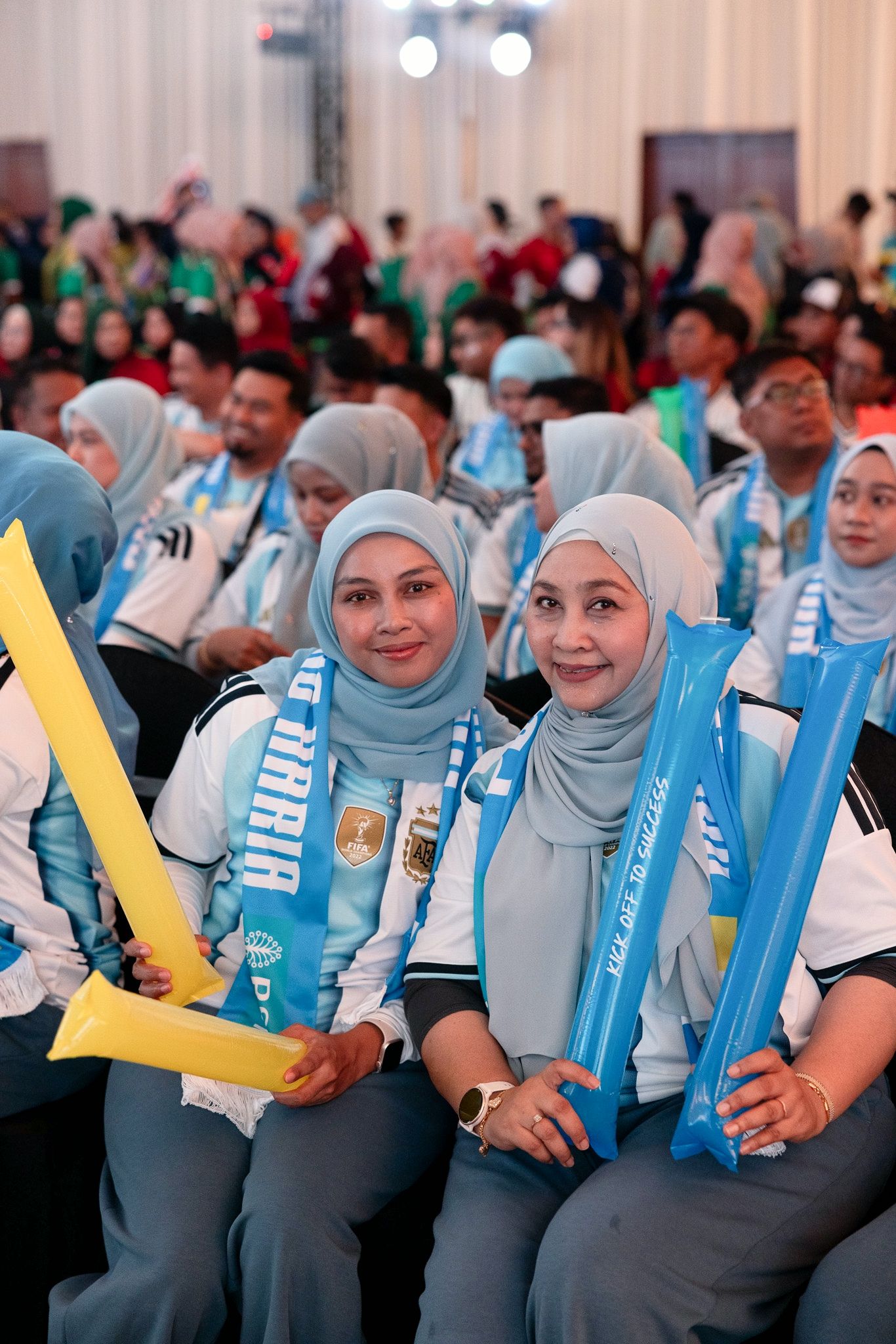 Two women in hijabs holding inflatable cheering sticks at a sports event, wearing light blue scarves and jerseys.
