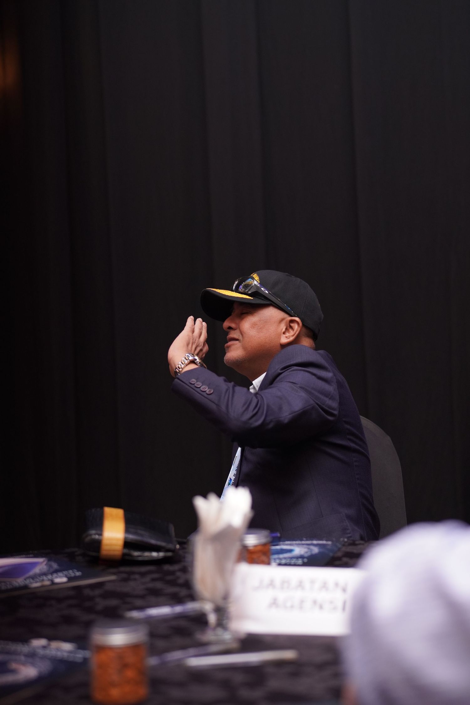 Business professional at conference panel wearing navy baseball cap gestures during presentation or discussion.