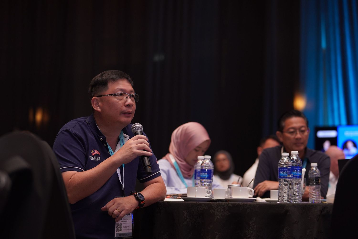 Business conference attendees seated at round tables in a dimly lit room with ambient blue lighting.