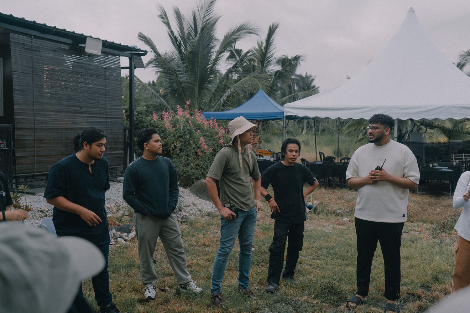 A group of people standing together on grass during an overcast day with palm trees in the background.