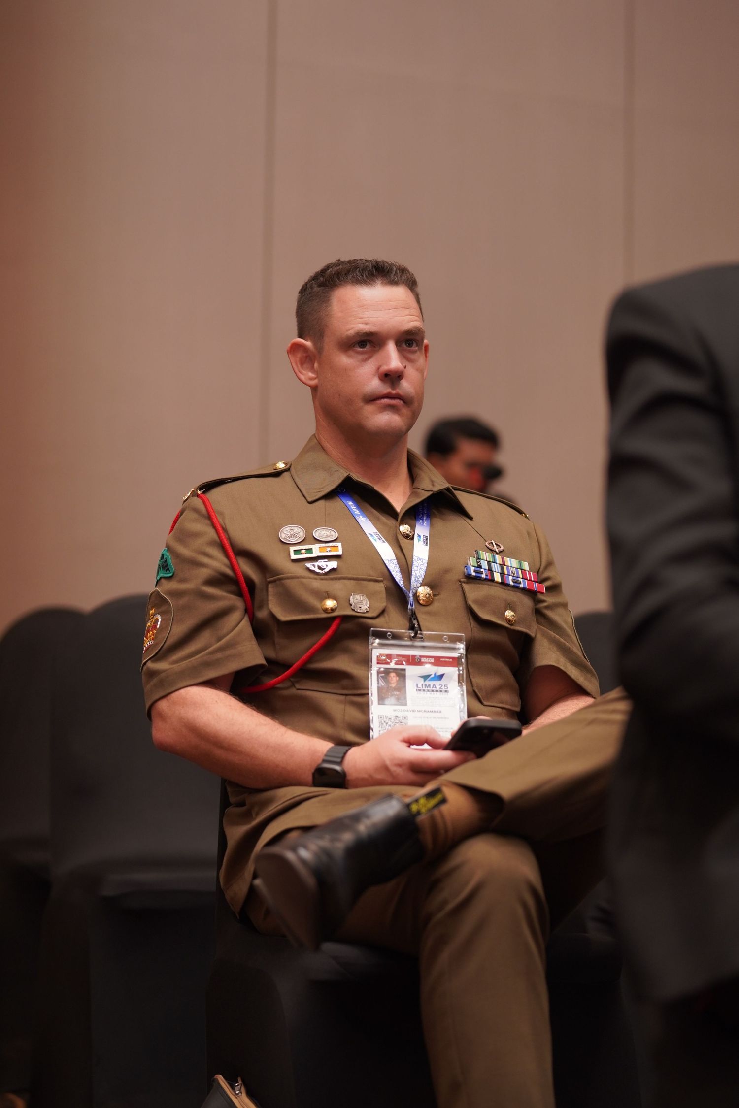 Military officer in brown uniform sits attentively during a presentation.
