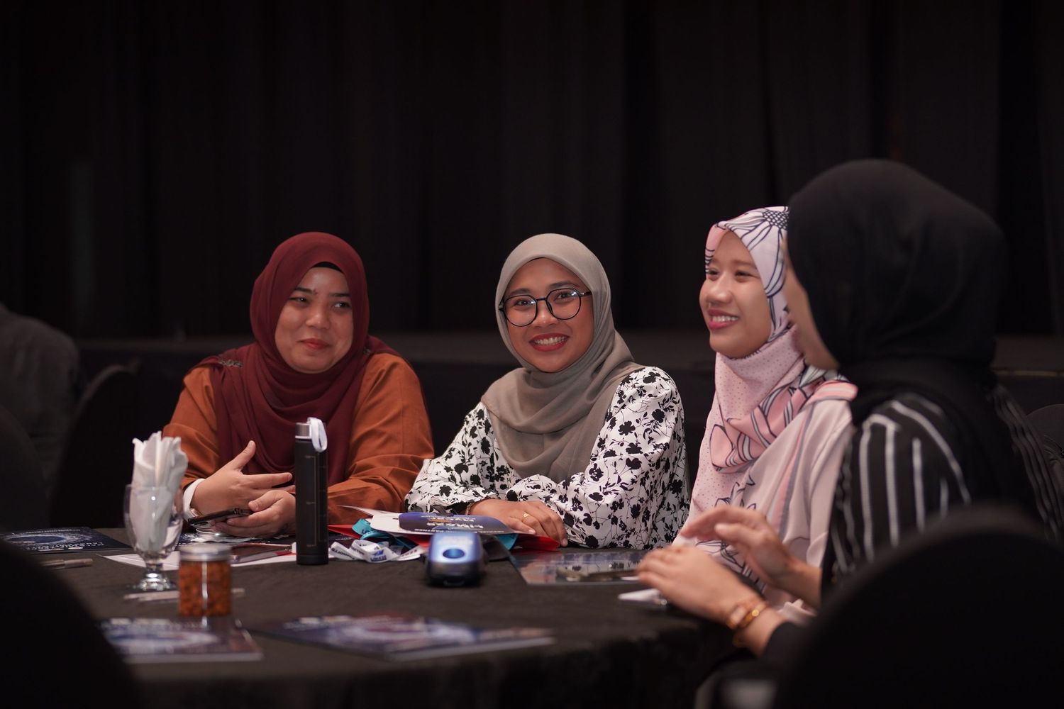 Three people in hijabs share a moment of laughter while seated at a table during what appears to be a conference.