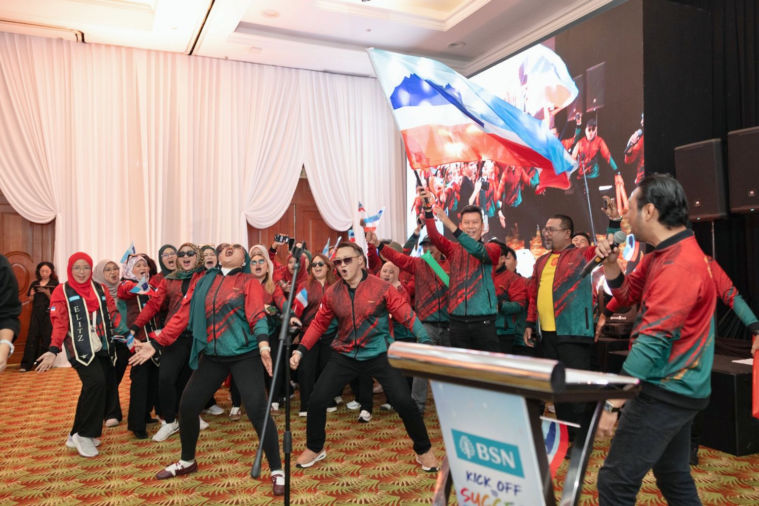 Group of people in red uniforms waving a flag at an indoor corporate event ceremony on a decorated stage.