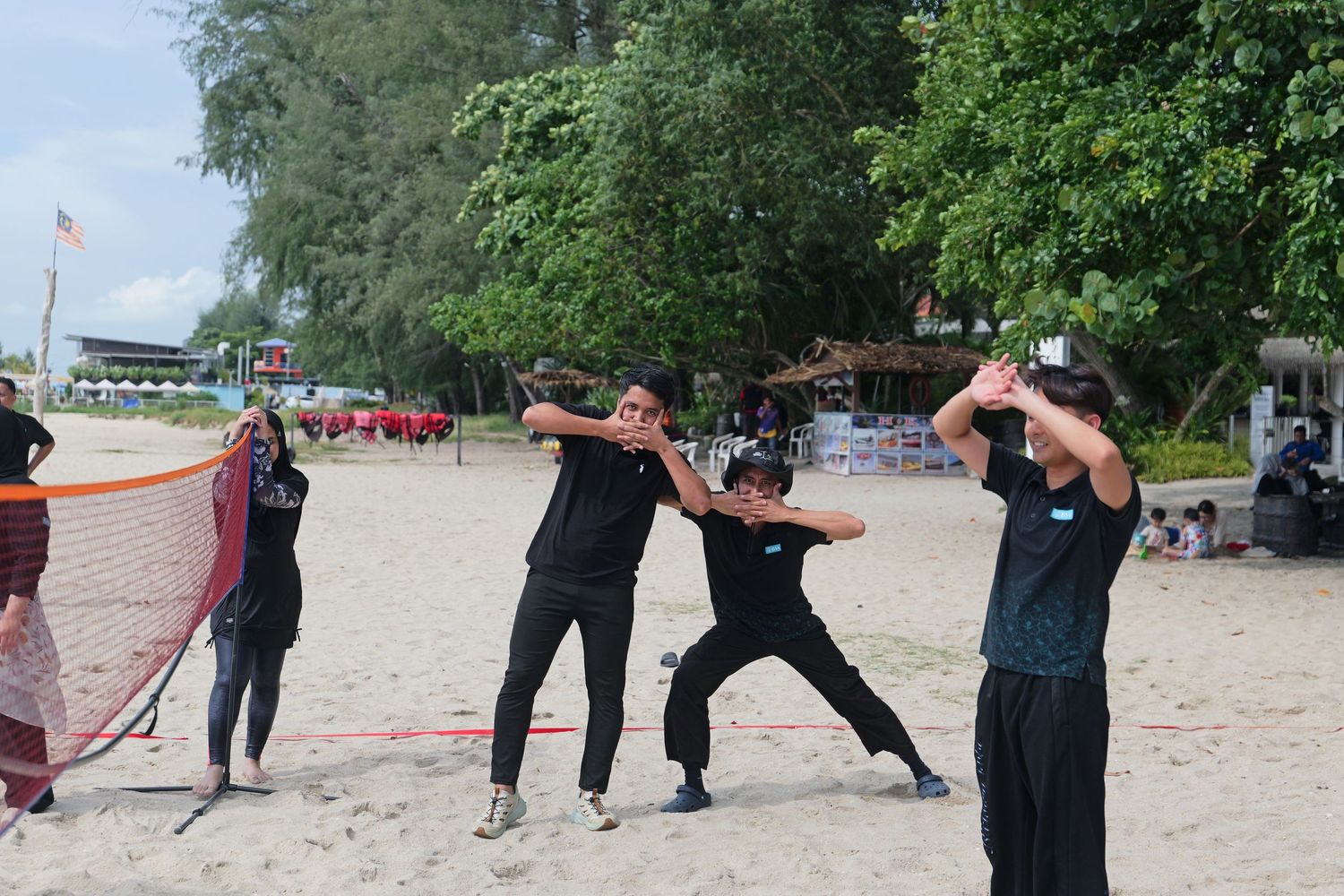 People in black clothing playing beach volleyball on sandy shore with trees in background.