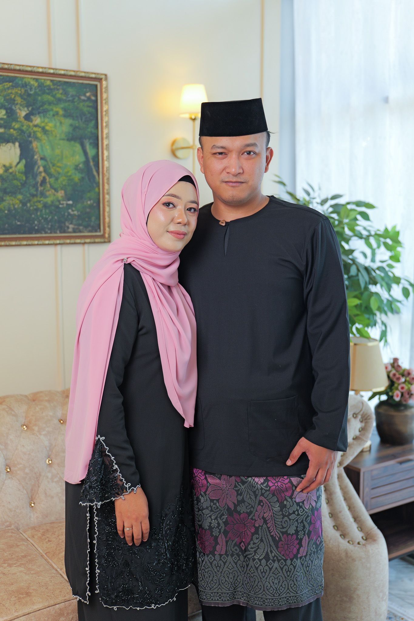 A couple in traditional Muslim attire poses together in a home setting with a pink hijab and black clothing.