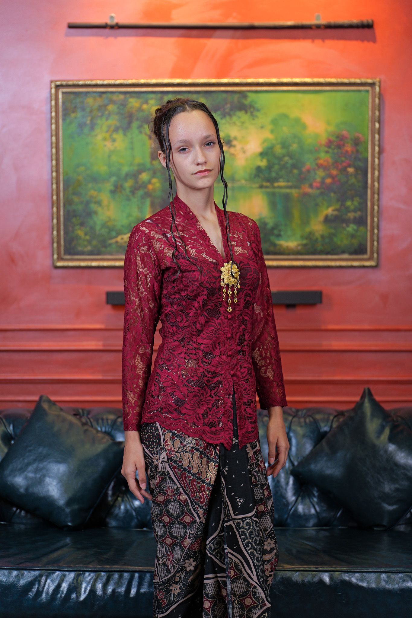 Traditional Indonesian fashion featuring a red lace kebaya top paired with batik sarong against a coral-colored wall with artwork.