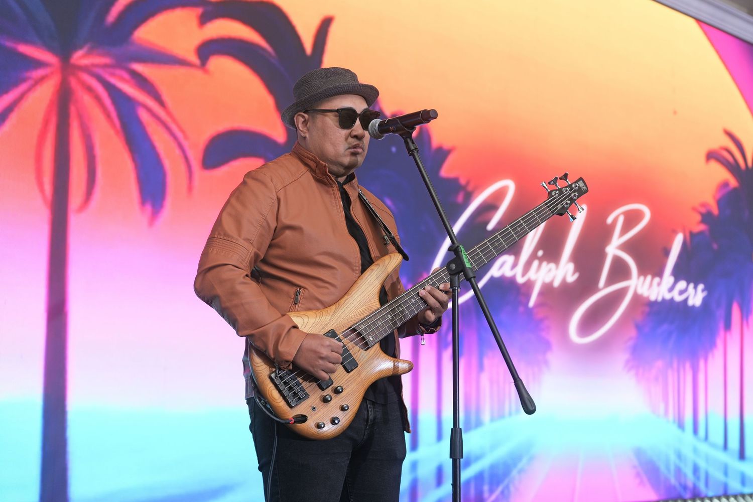 Male guitarist performing on stage at a Caliph Buskers concert with colorful tropical sunset backdrop.
