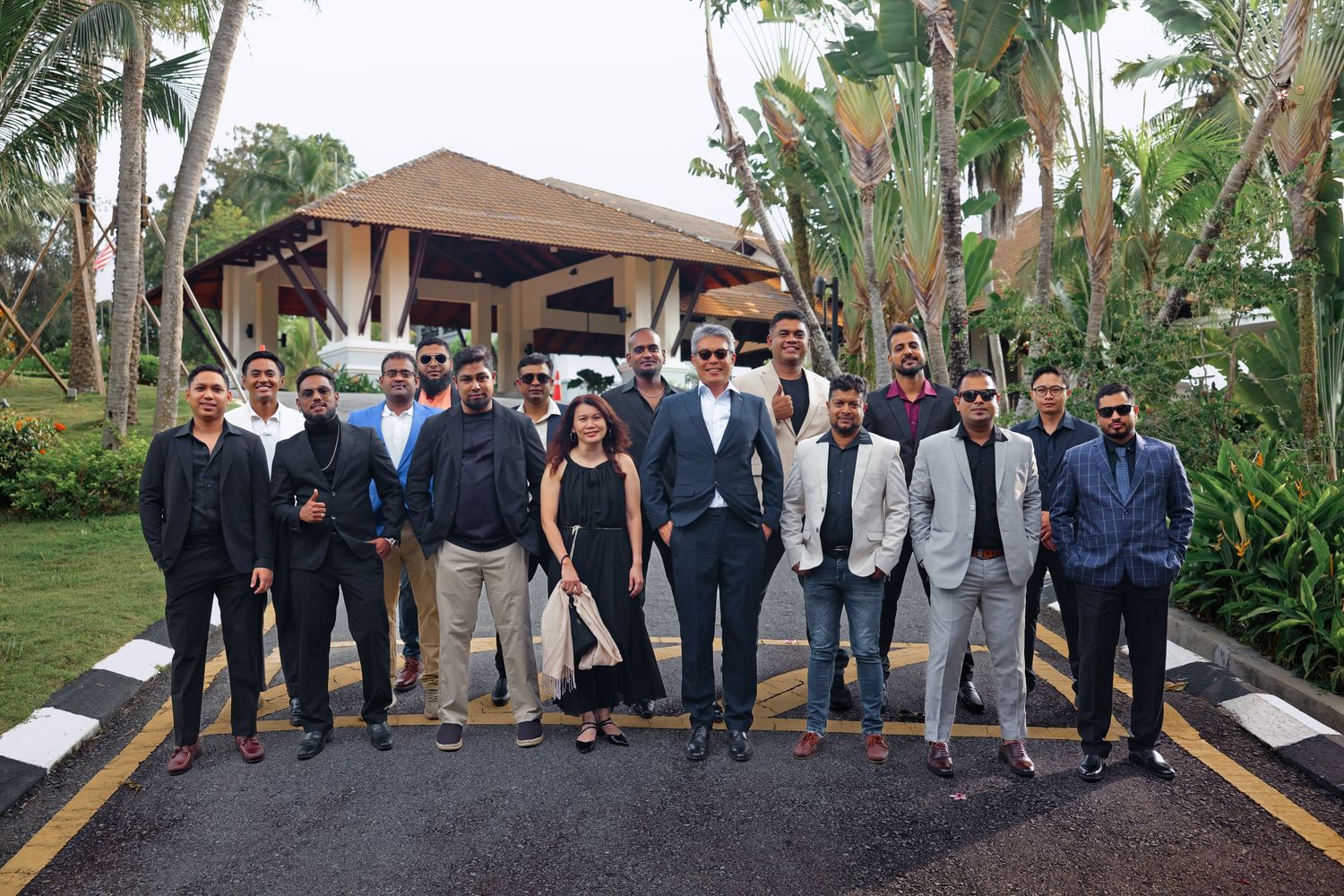 A large group of business professionals pose outdoors at a tropical hotel entrance for a group photo.