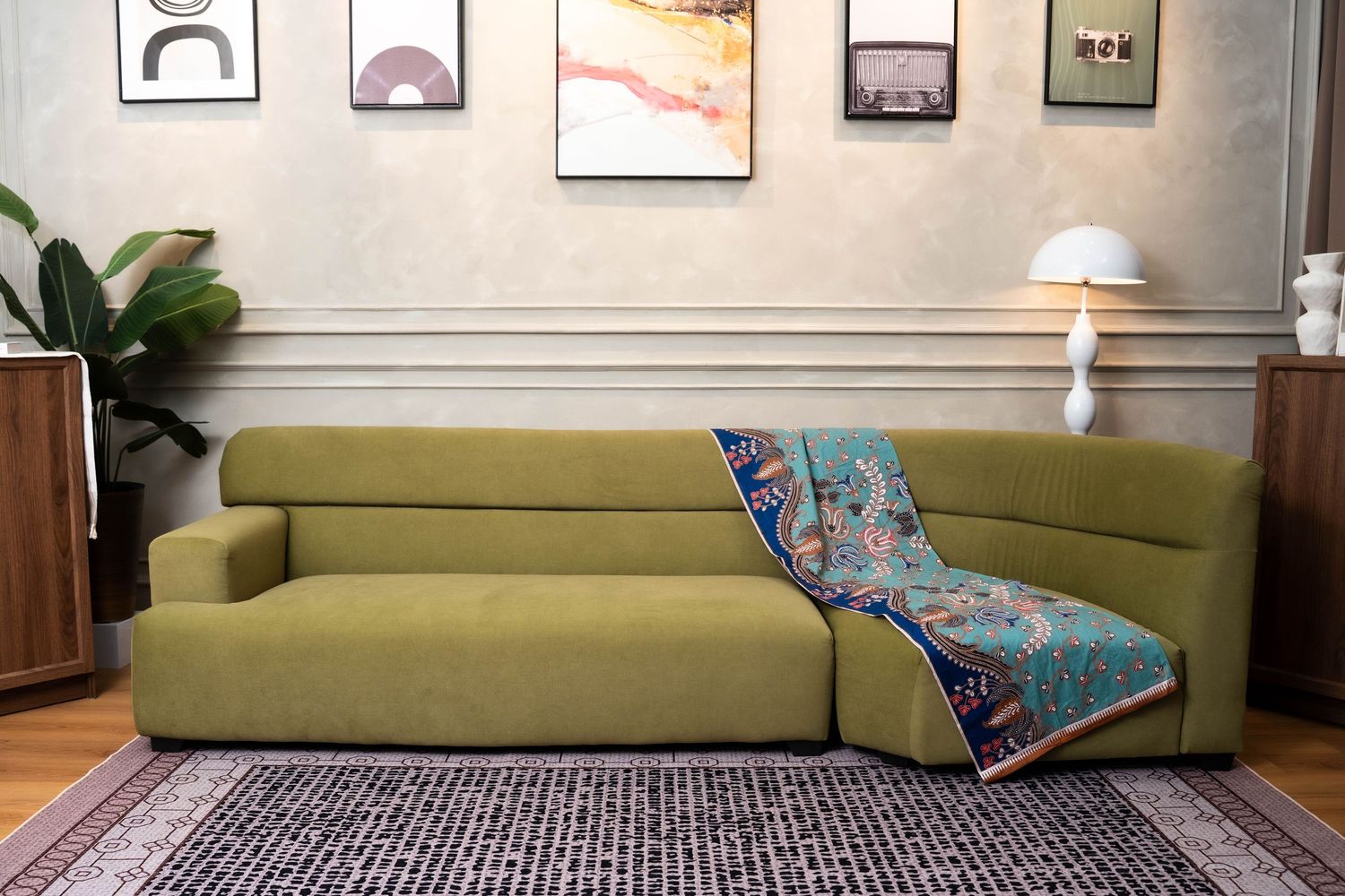 Modern olive green sectional sofa with colorful throw blanket in living room with wall art and table lamp.