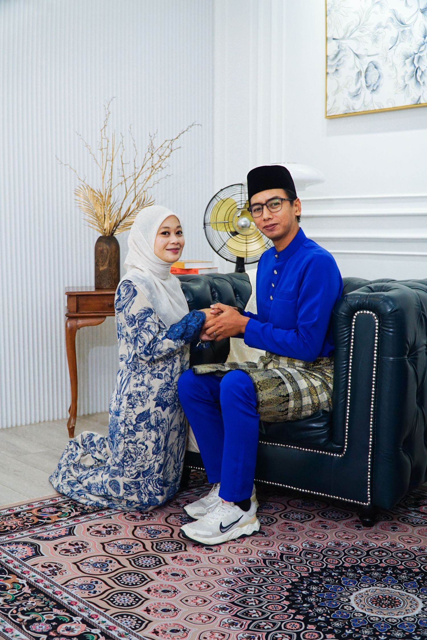 Couple in traditional attire sitting on patterned carpet in living room with decorative white walls.
