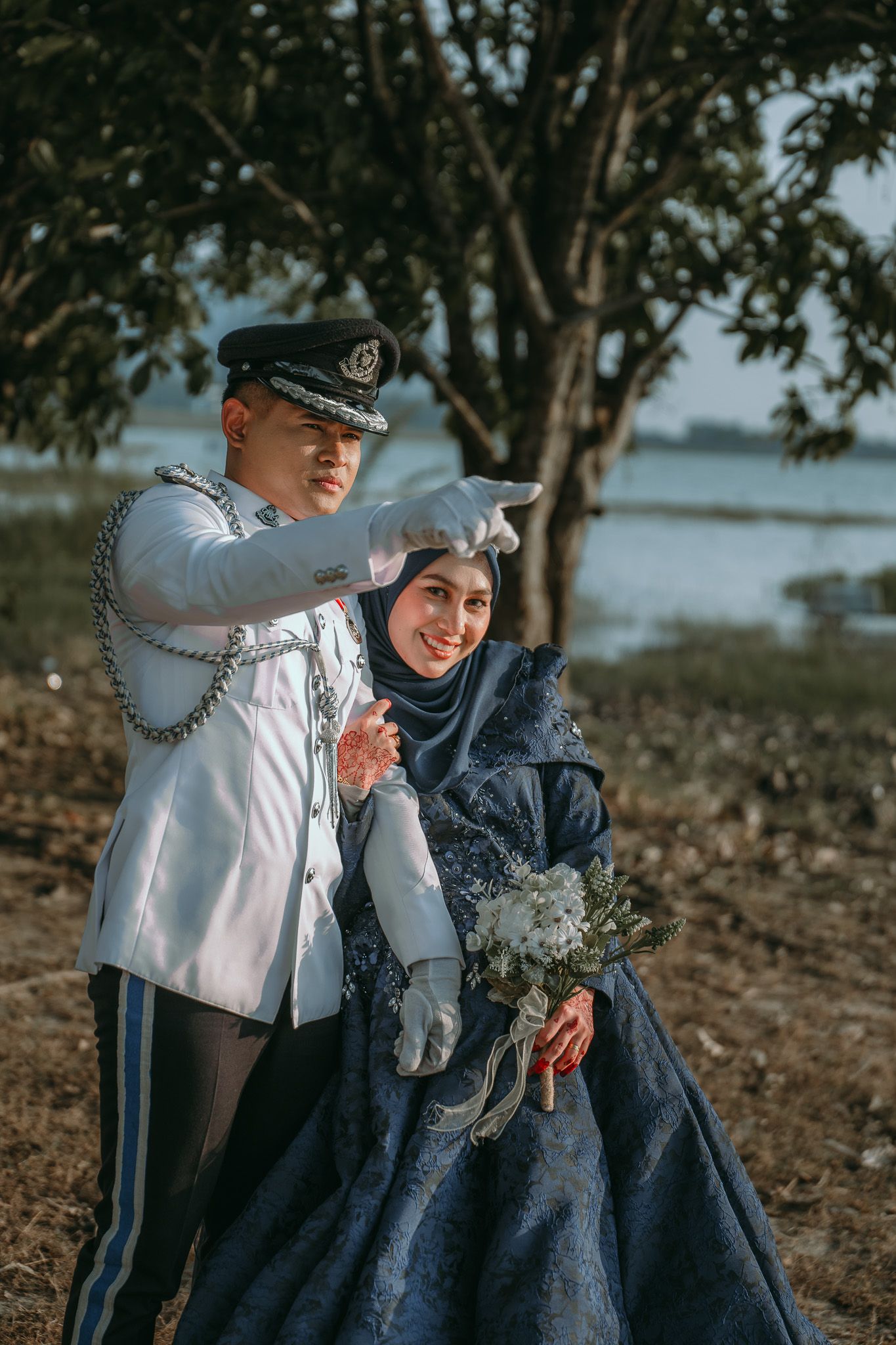 Military officer and woman in traditional hijab pose together on lakeshore with bouquet.