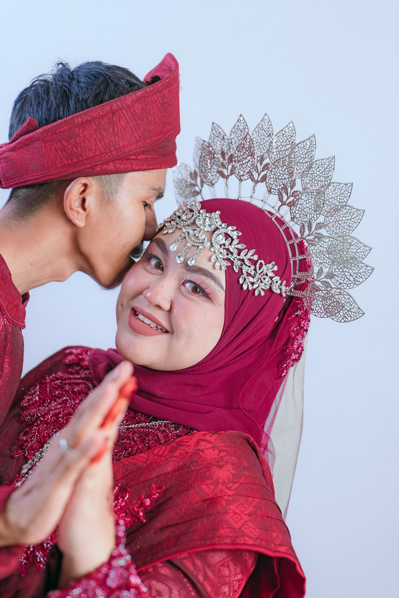 Traditional Indonesian wedding couple in red attire with bride wearing ornate gold crown and hijab during intimate moment.