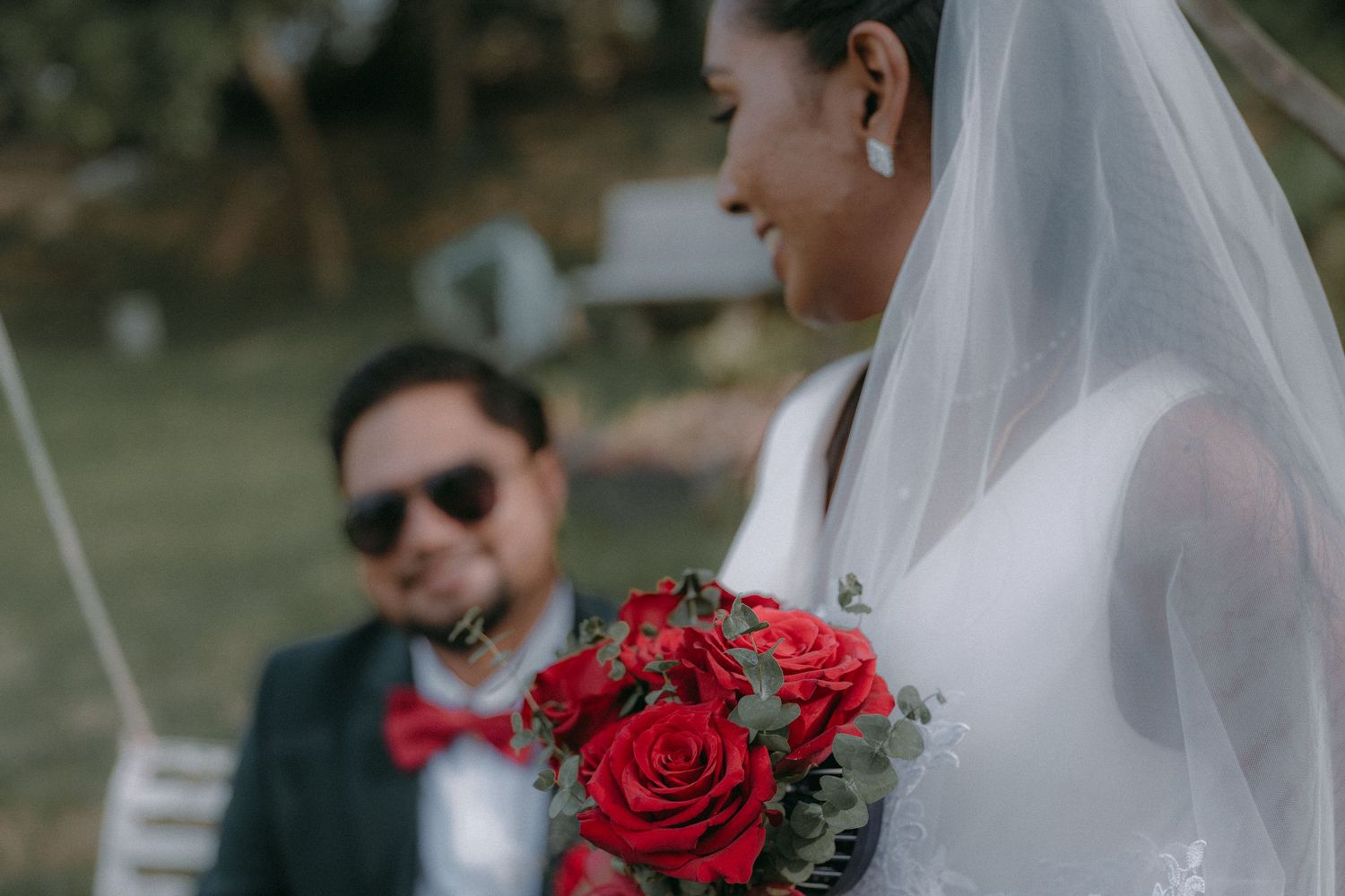 Bride in white dress with red rose bouquet stands with groom in outdoor ceremony.