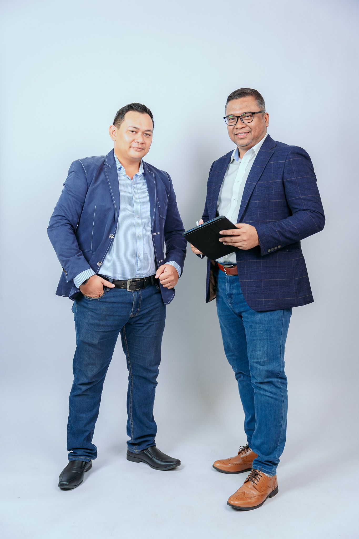 Two businesspeople in navy suits and jeans examine mobile devices against a light gray studio background.