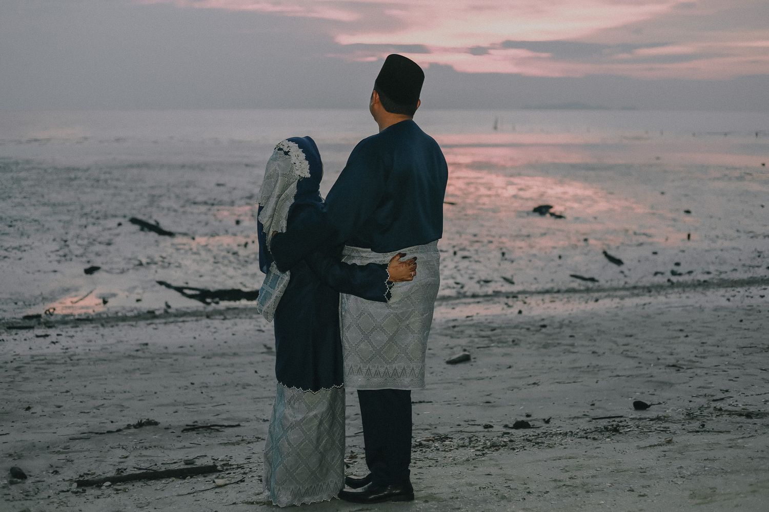 A couple standing together on a beach at sunset, looking out at the misty horizon under pink clouds.