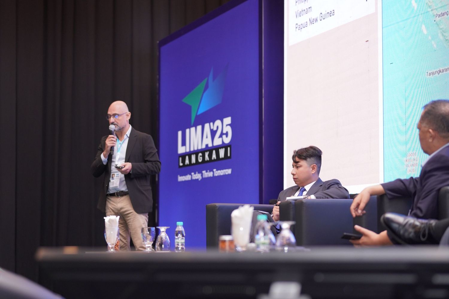 Speaker presenting at LIMA25 conference with panelists seated at table.