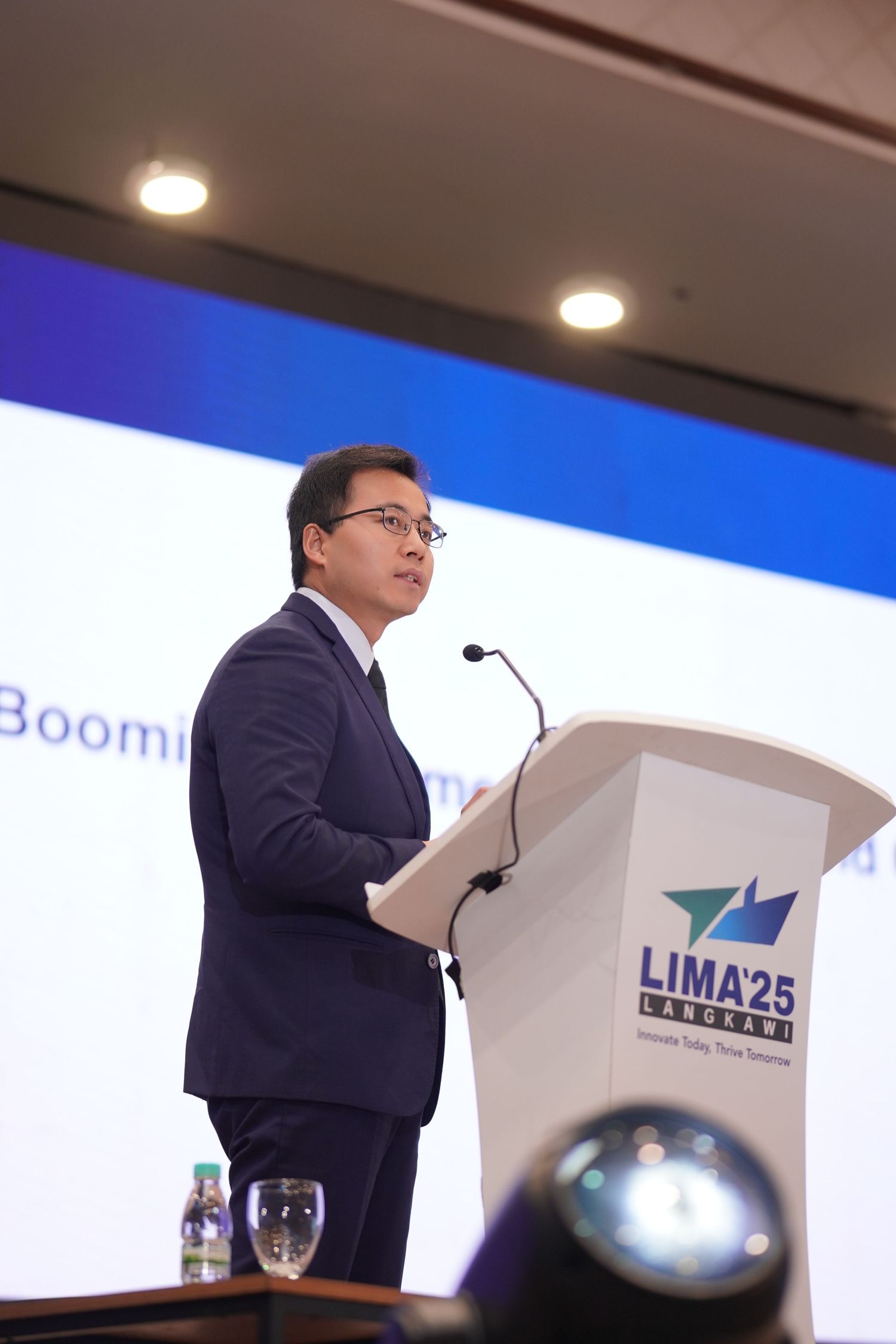 A speaker in a navy suit stands at a podium with the LIMA25 logo during a conference presentation.