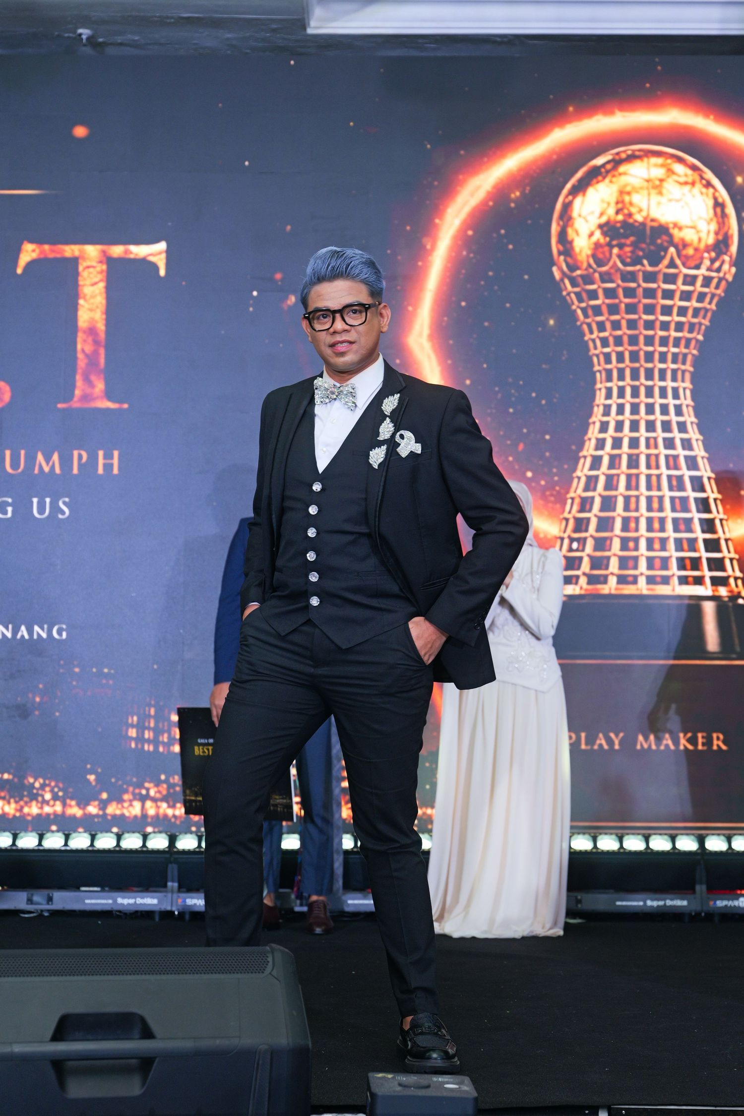 Man in black suit with blue hair and bow tie posing at awards ceremony.