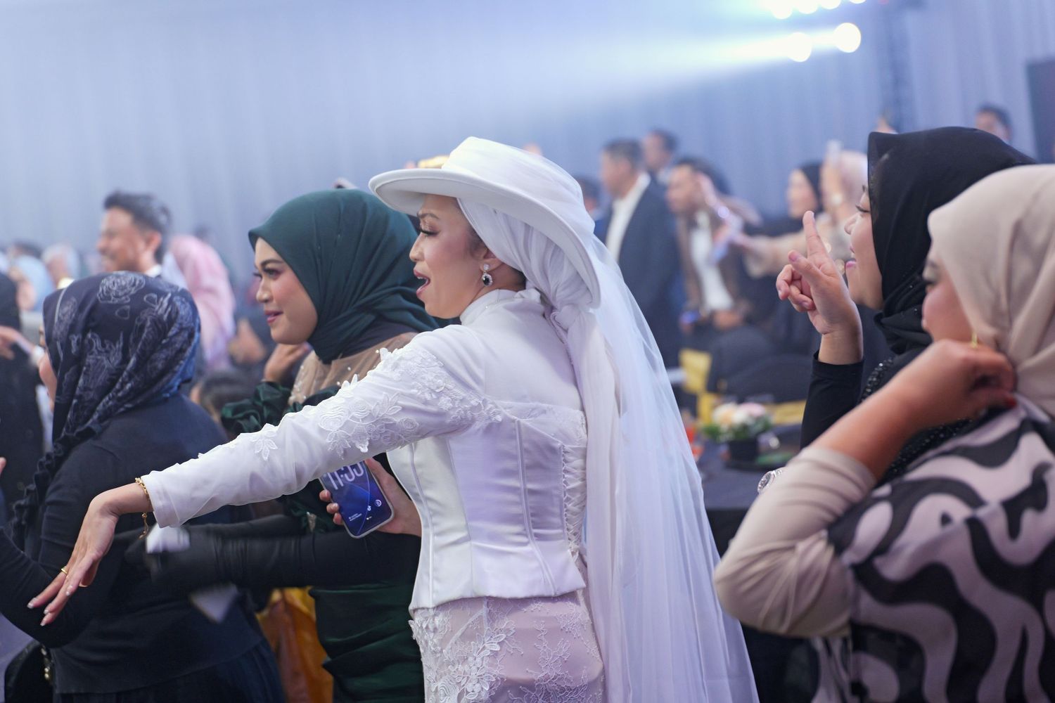 Woman in white hat and kebaya dancing joyfully surrounded by guests at a formal celebration.