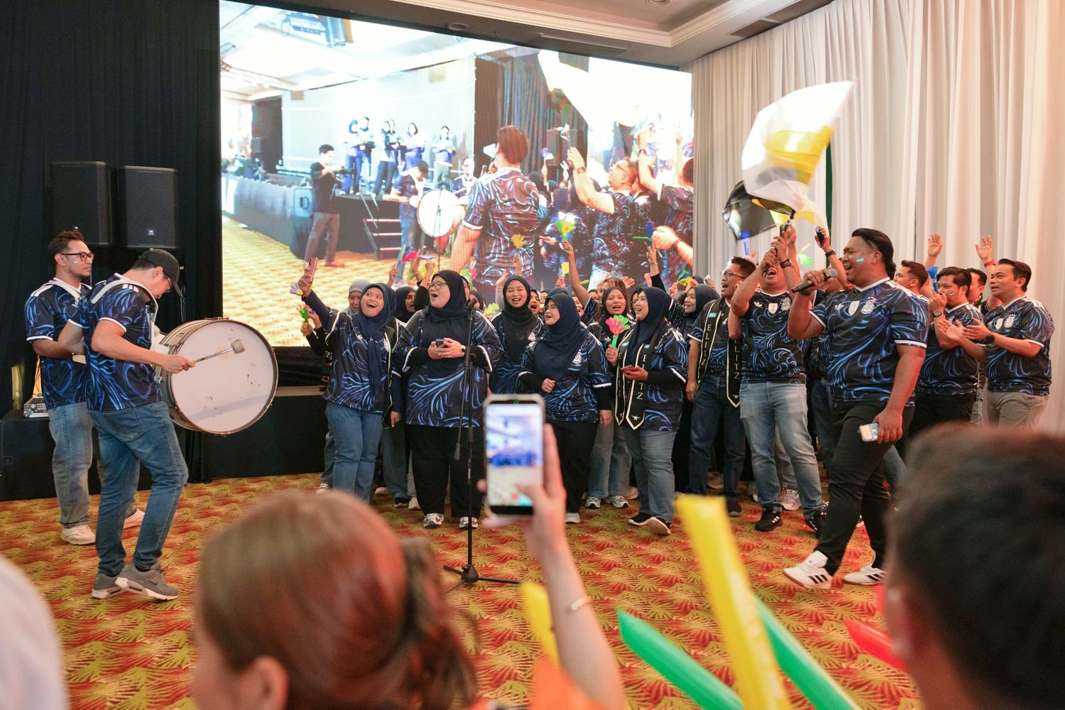 A lively corporate event features a marching band performing indoors with drums and instruments for an enthusiastic audience.