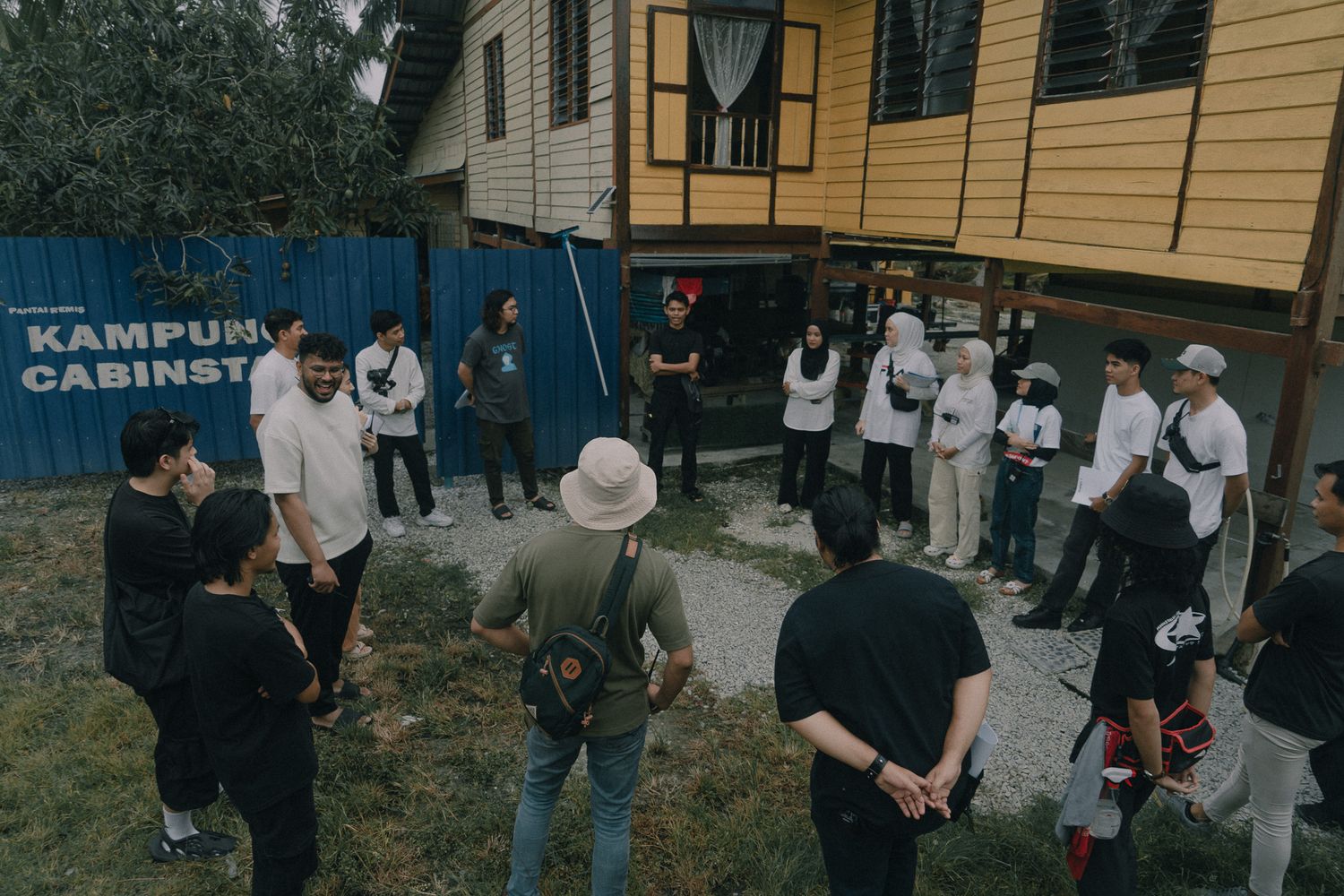 A group of people gathered outside a yellow building with a blue Kampung Cashney sign in a casual outdoor setting.