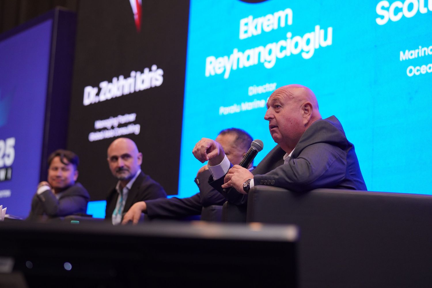 Professional conference stage with digital name displays and speakers panel illuminated in blue.