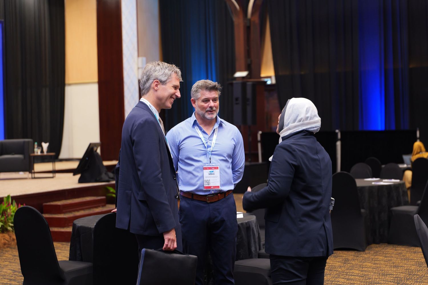 Business professionals engaged in discussion at a conference event with stage and lighting in background.