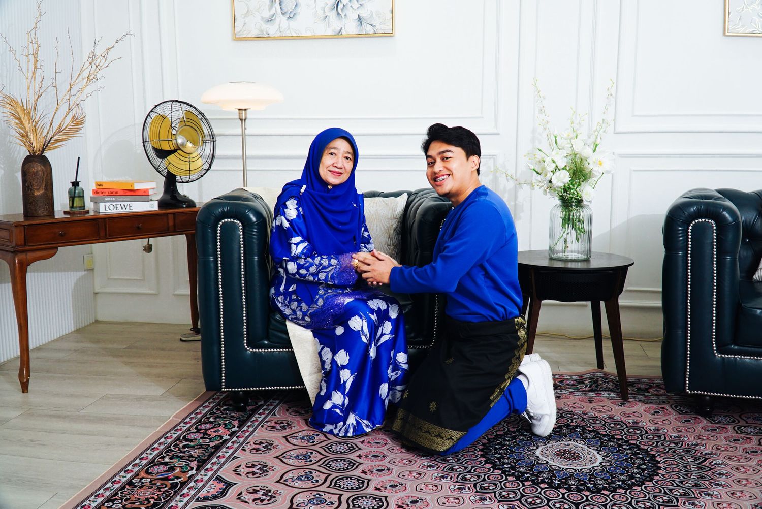 Two people in blue outfits sit on a patterned rug in a bright white room with modern decor.