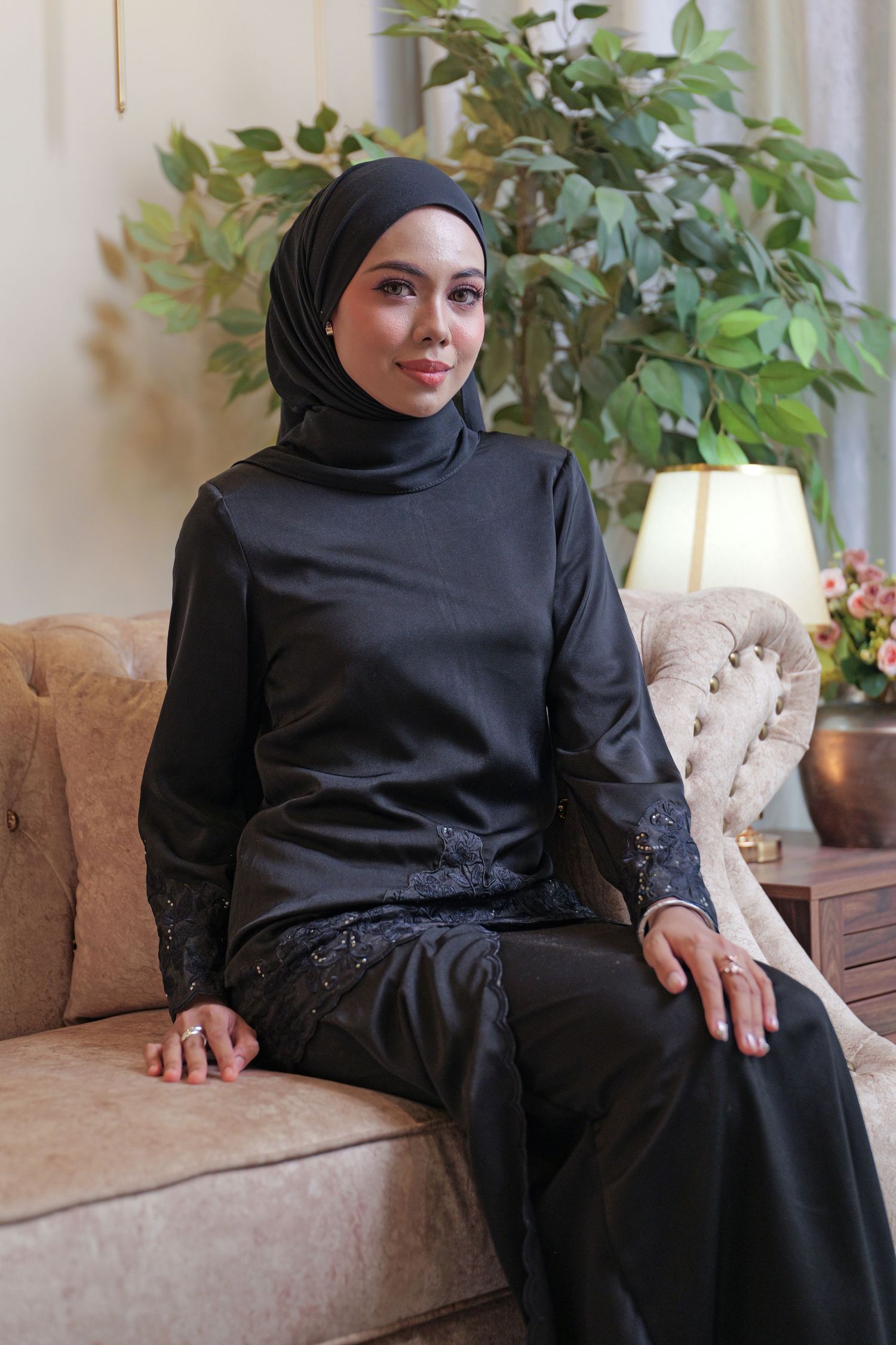 A person wearing a black abaya and hijab sits elegantly on a beige couch with plants in the background.