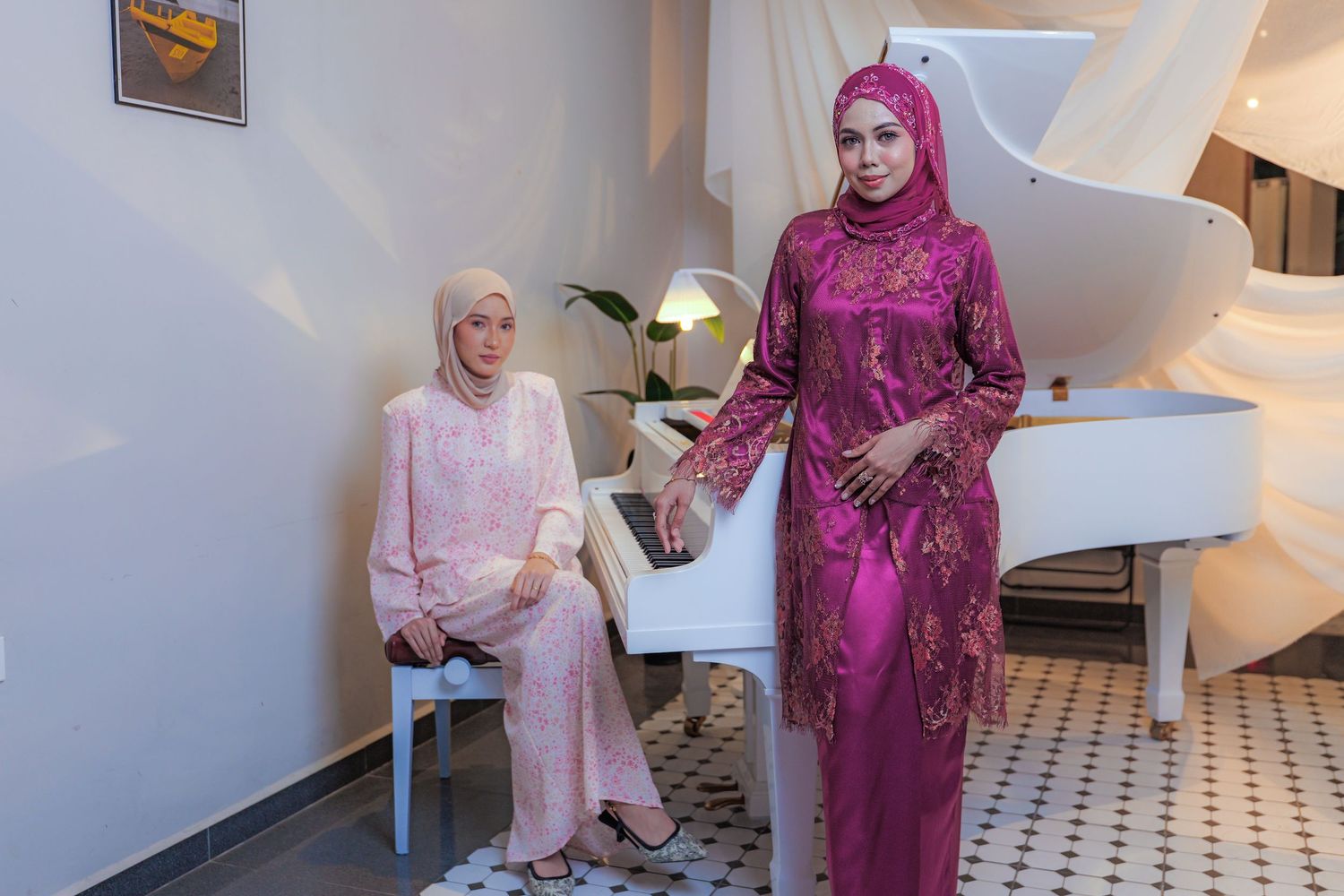 Two people in elegant dresses sit together at a white piano in a bright room with tiled flooring.