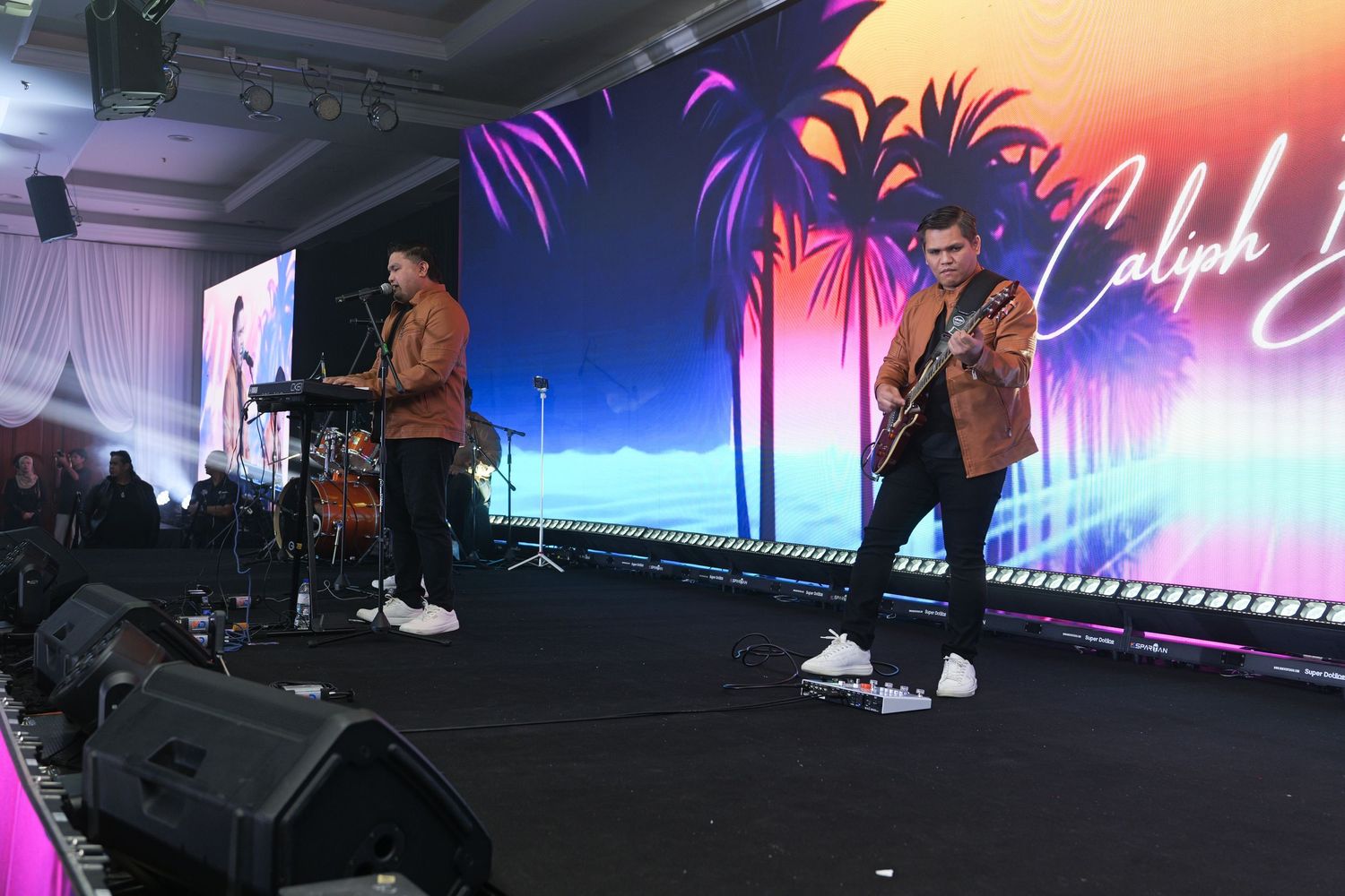 Two musicians performing on stage with colorful tropical-themed backdrop at an indoor entertainment event.