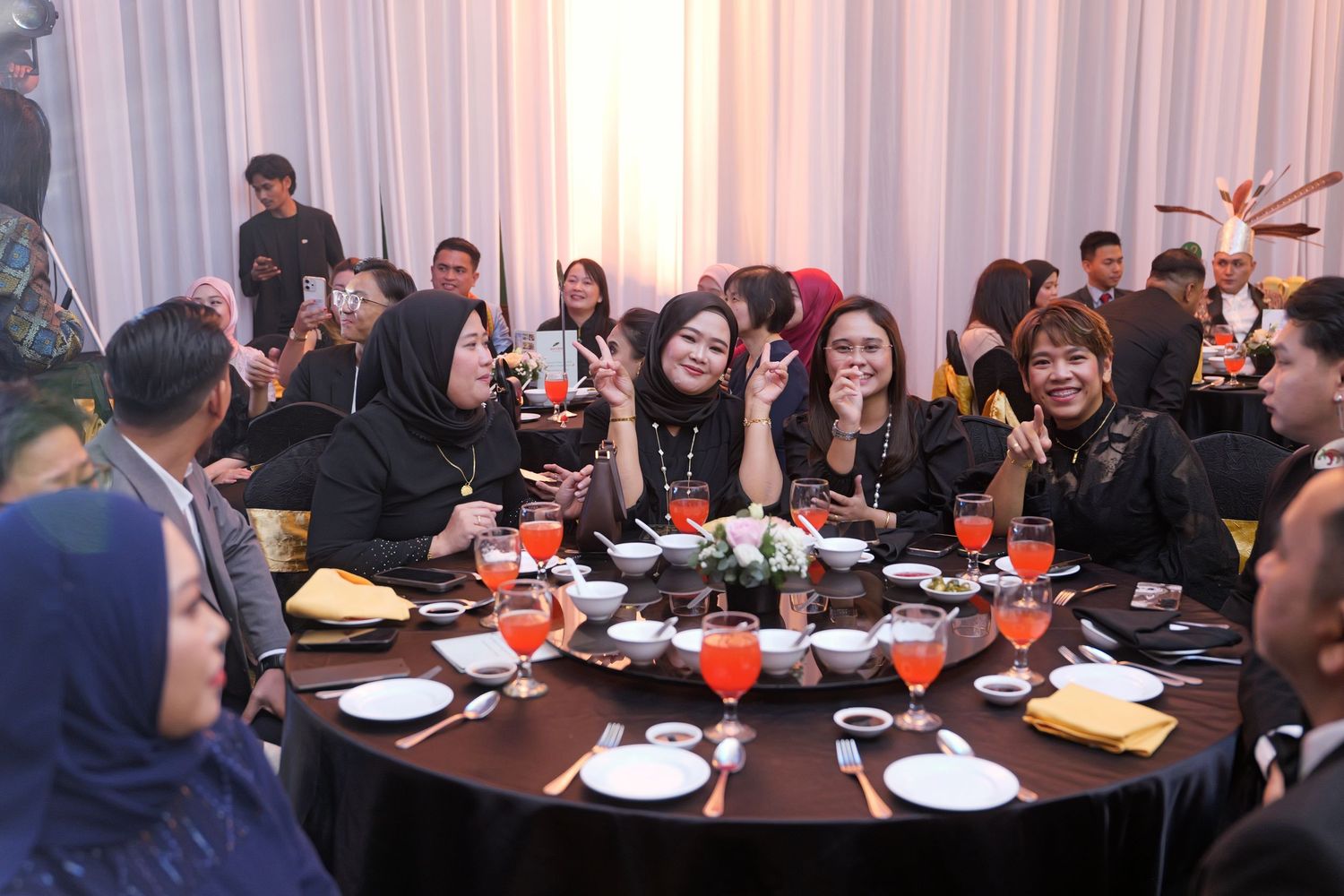 Group of elegantly dressed guests enjoying a formal dinner banquet, seated at a round table with orange drinks.